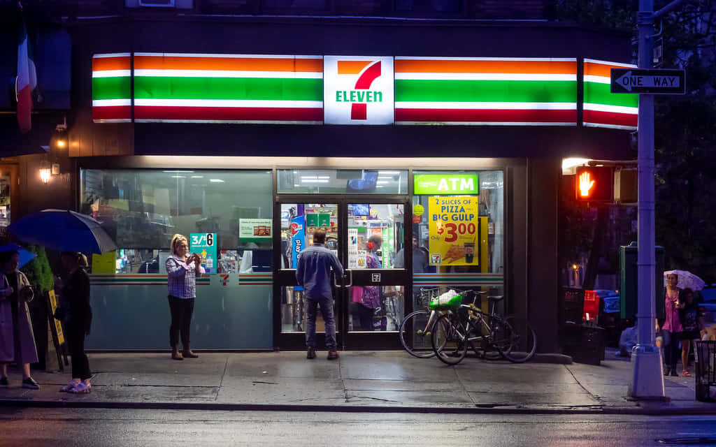 7-Eleven，隨時滿足您的零食需求 壁紙 — 一個雨夜場景，在一間燈火通明的7-Eleven商店外，有人撐著雨傘，附近停著一輛自行車。