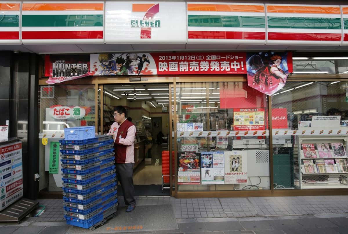 男子在日本便利商店外堆疊藍色板條箱，商店貼有動畫海報。