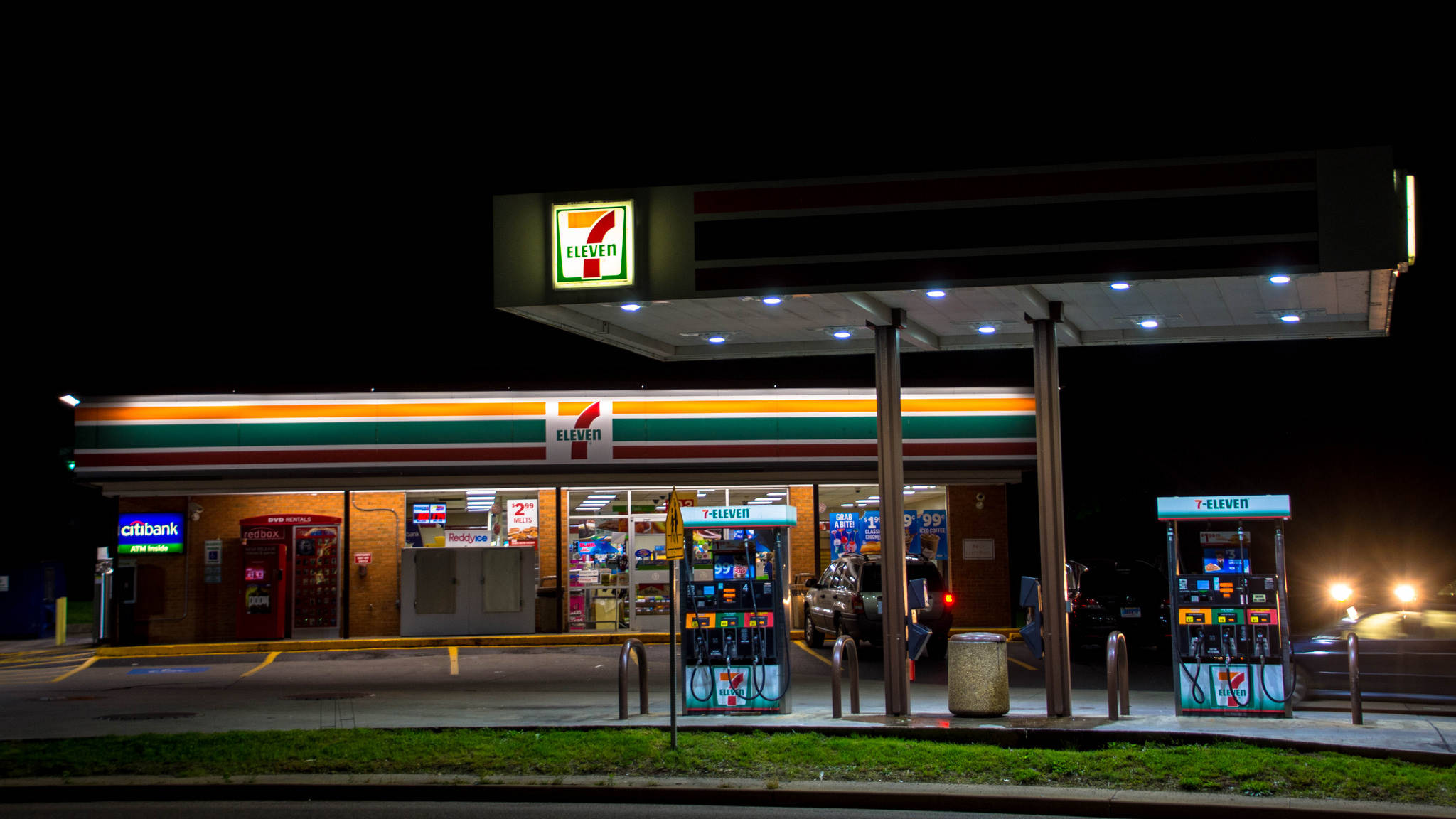 7-Eleven加油站 壁紙 — 夜晚明亮照明的7-Eleven加油站，發光的油泵和反射霓虹燈牌的商店櫥窗。