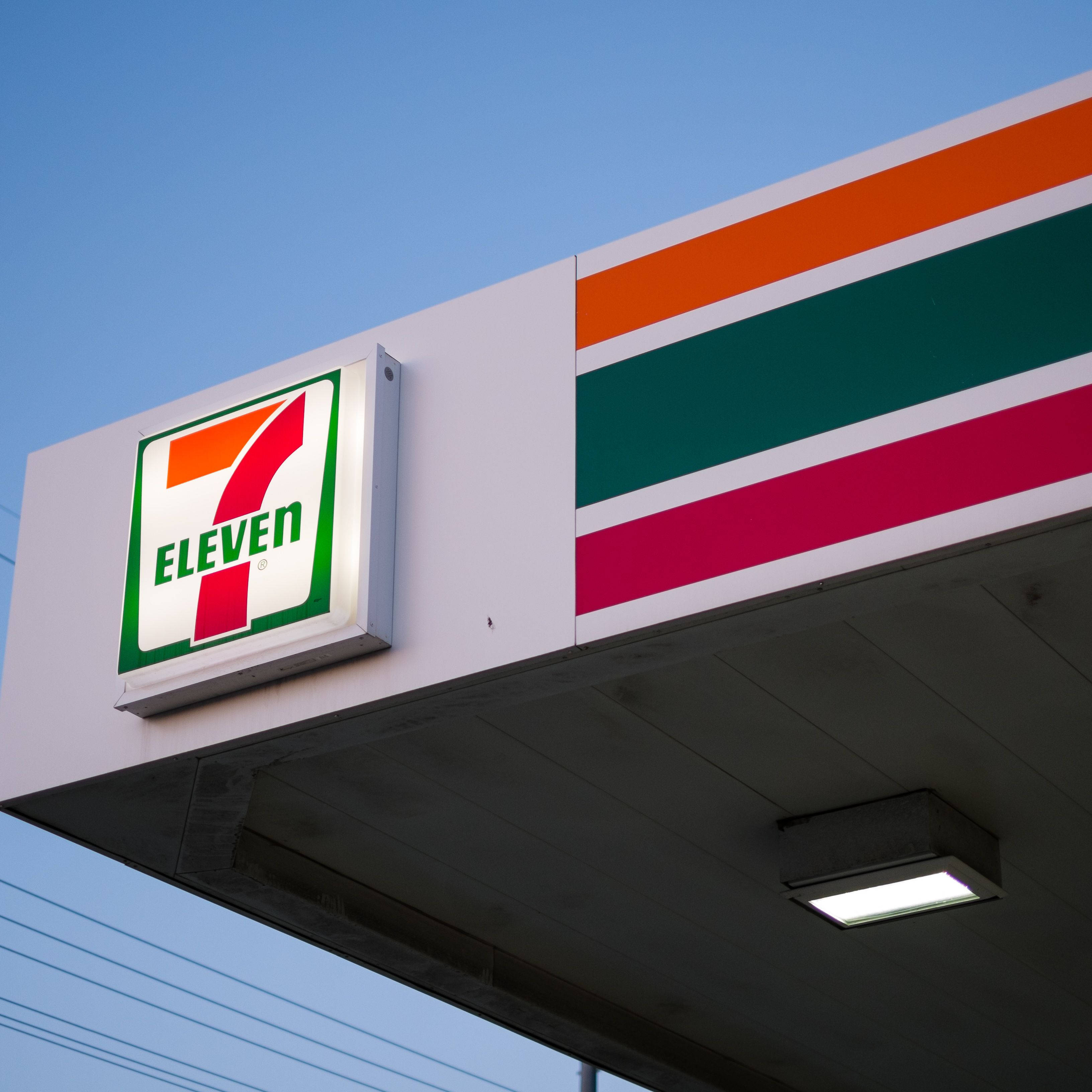 7-Eleven招牌 壁紙 — 一個明亮照明的7-Eleven招牌，帶有醒目的橙色、綠色和粉紅色條紋，背景是清澈的藍天。