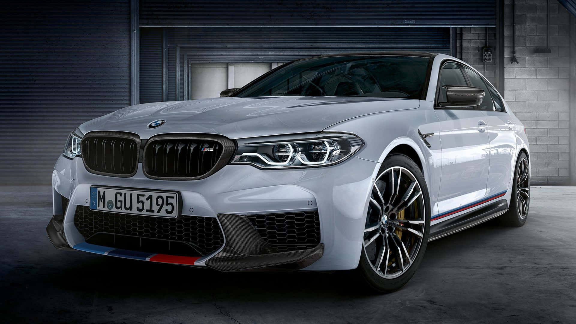 720p M 系列背景：白色 BMW 2 系列室內拍攝 壁紙 — 一輛時尚的銀色BMW M5轎車，帶有紅、藍、白賽車條紋，停在光線昏暗的工業車庫中。
