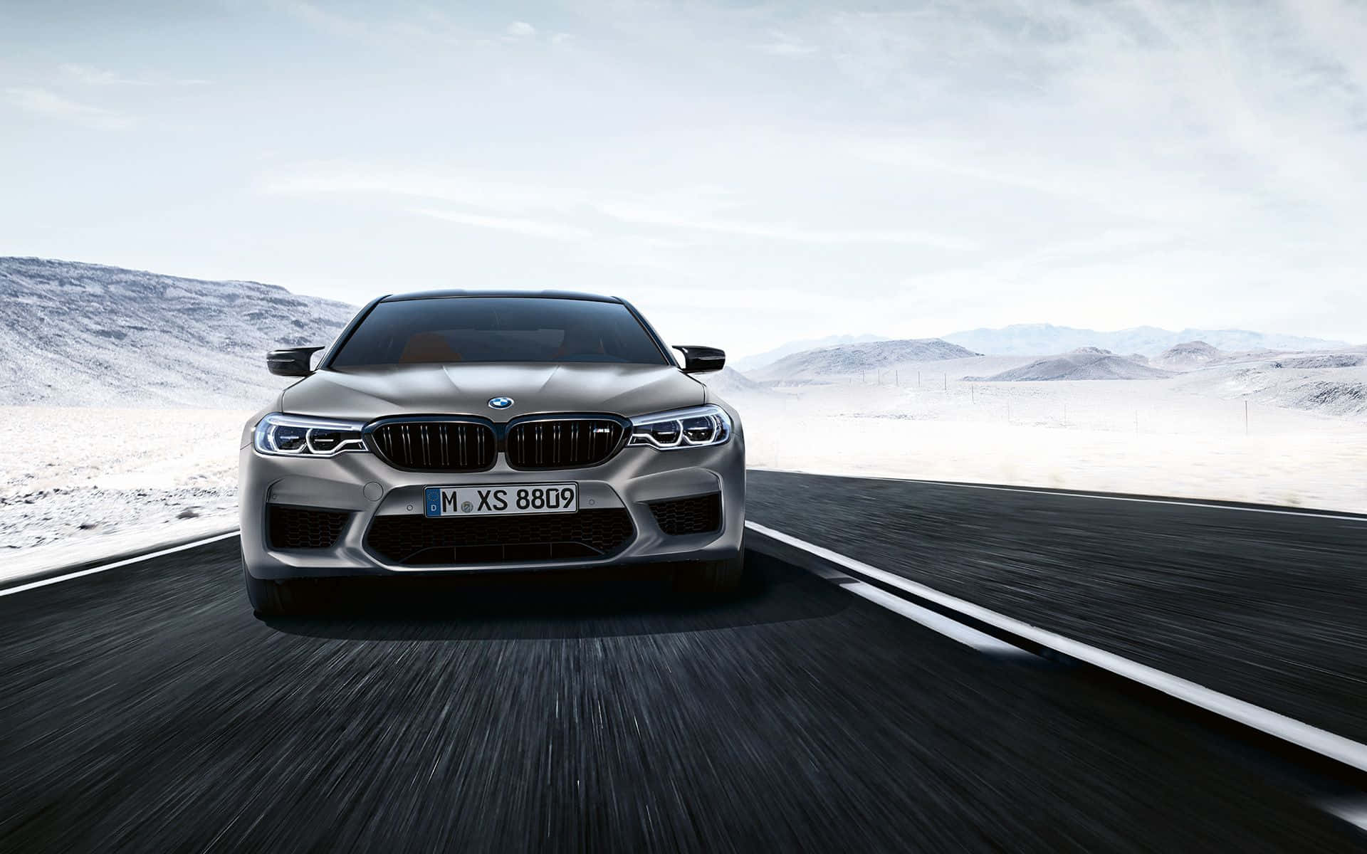 銀色BMW M5在日落時分於沙漠公路上奔馳，背景是雪山。