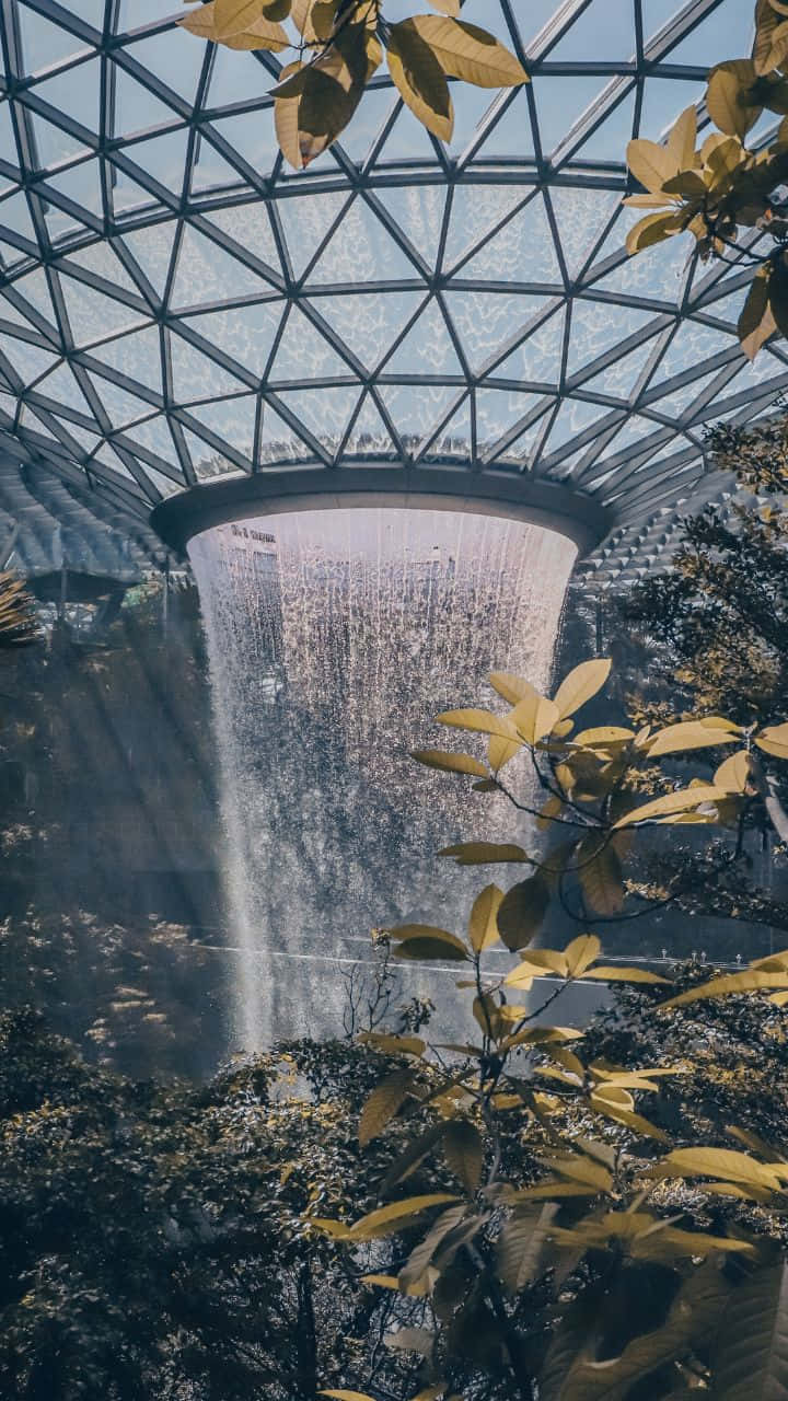 720p 旅行背景：新加坡 The Jewel 壁紙 — 一個宏偉的室內瀑布在幾何玻璃穹頂下傾瀉而下，周圍環繞著金色的秋葉。