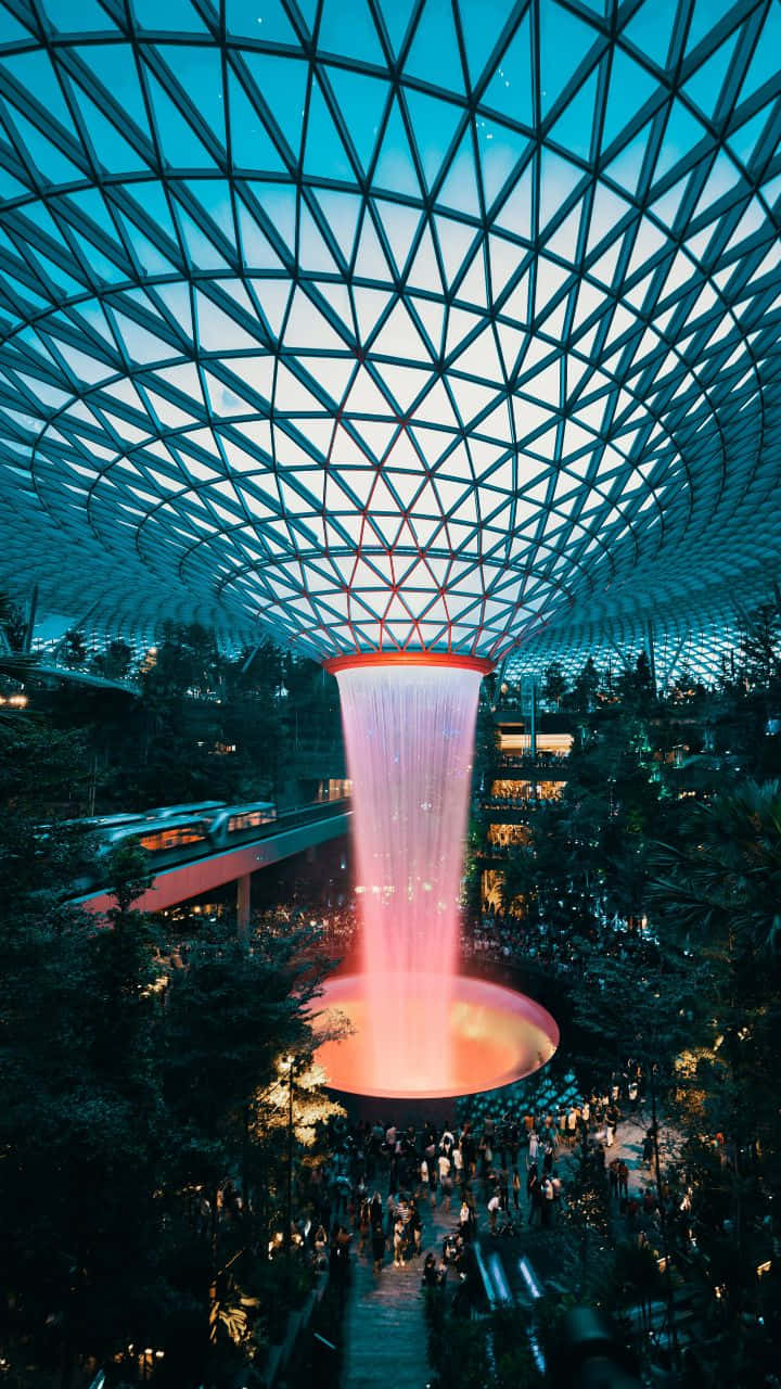 720p 旅行背景：新加坡樟宜機場 The Jewel 壁紙 — 在黃昏時分，一個發光的粉紅色瀑布從幾何玻璃圓頂傾瀉而下，落在繁忙的室內花園上方。