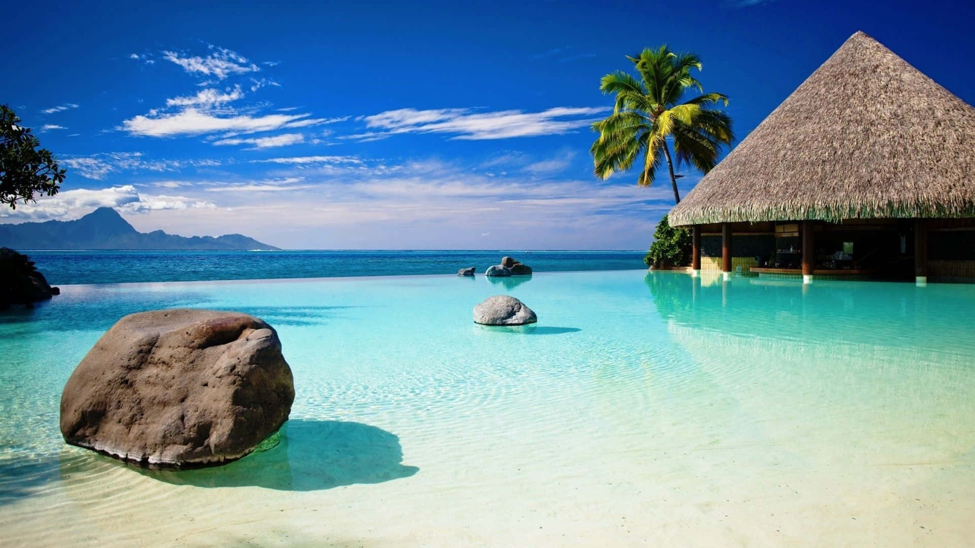 720p 海灘巨岩小屋背景 壁紙 — 一個寧靜的無邊際泳池與碧綠海洋融為一體，旁邊是茅草屋頂別墅，在明亮的藍天下有零星雲朵。
