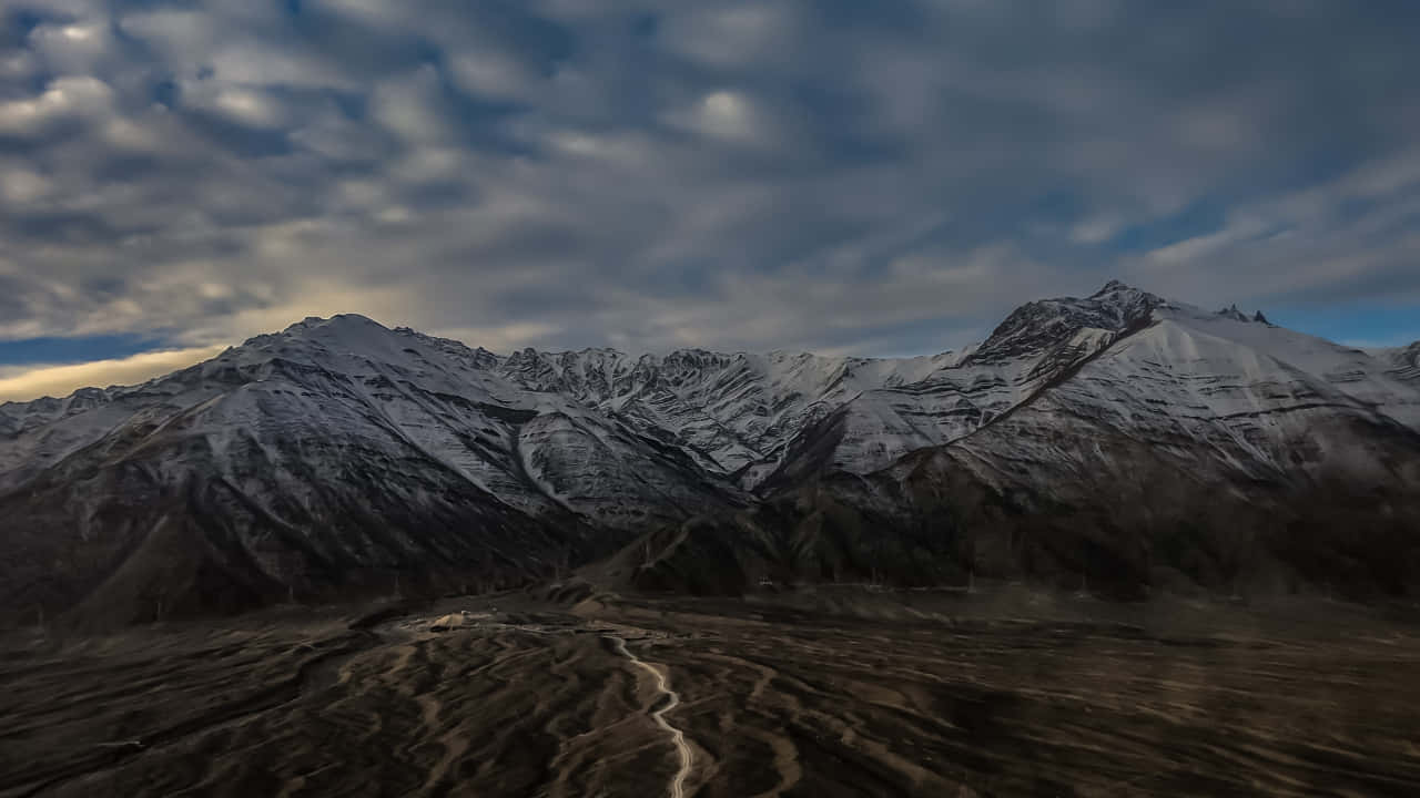 720p 自然雪山雲霧背景 壁紙 — 一條蜿蜒的泥土路穿過廣闊貧瘠的山谷，下方是雄偉的雪山，上方是戲劇性的多雲天空。
