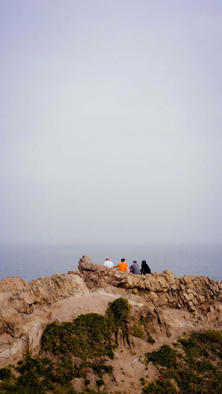720p旅行背景 - 天空與風景的完美融合 壁紙 — 四個人坐在崎嶇的懸崖邊緣，俯瞰著朦朧天空下平靜的海面。