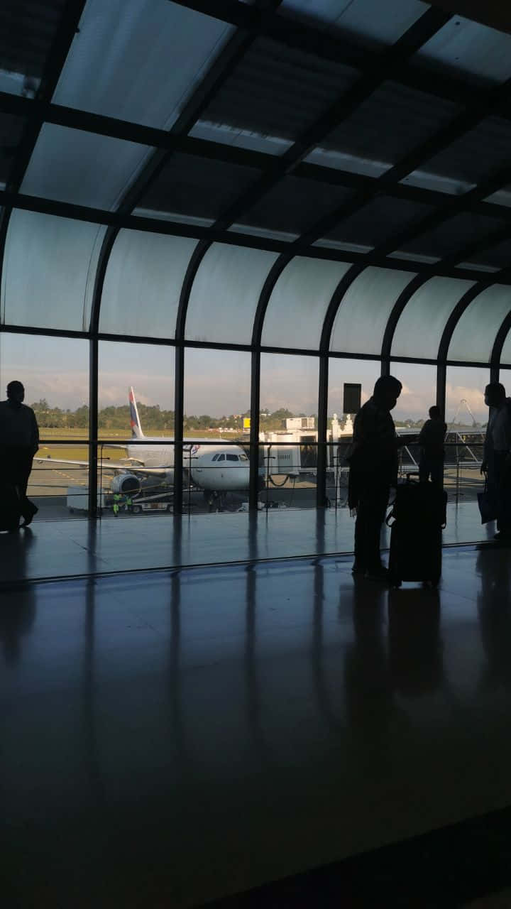 720p旅行背景：機場乘客登機閘口 壁紙 — 剪影的旅客帶著行李站在現代機場航站樓內，透過彎曲的玻璃窗凝視著停放的飛機。