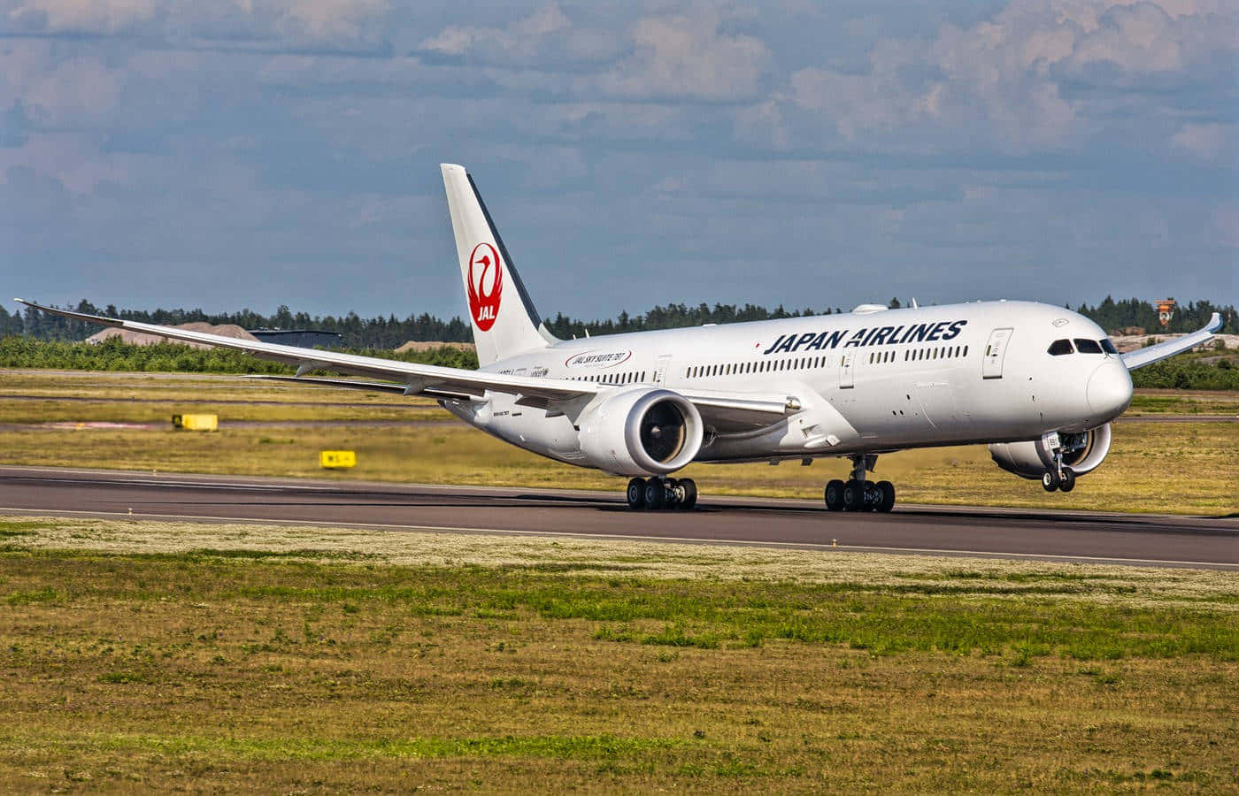 日本航空波音787起飛 壁紙 — 一架日本航空的波音787夢幻客機在部分多雲的天空下，於跑道滑行，周圍有綠草和遠處的樹木。