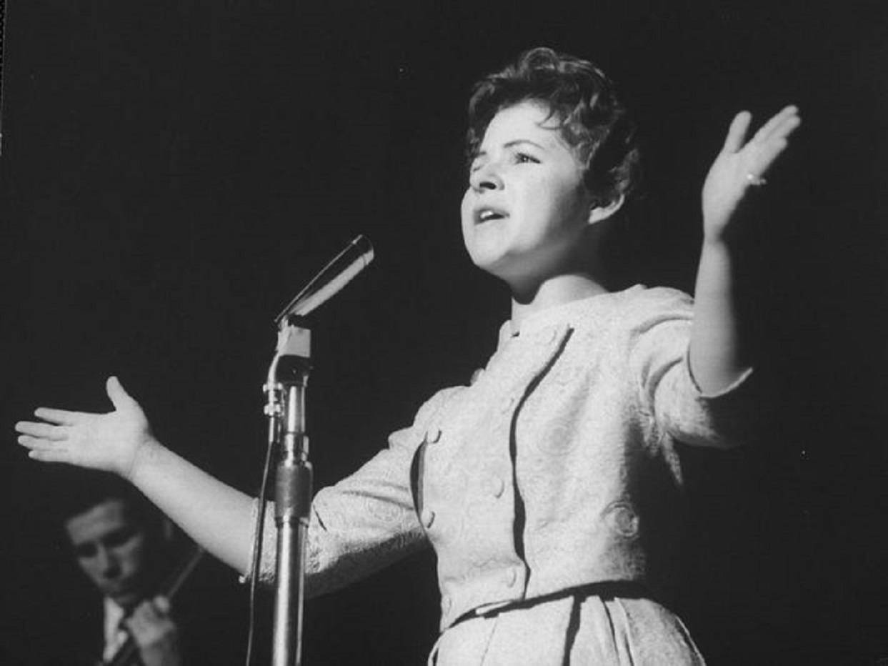 80年代復古流行歌手Brenda Lee舞台演出 壁紙 — 一張復古黑白照片，一位女性在昏暗的舞台上熱情地張開雙臂對著麥克風唱歌。