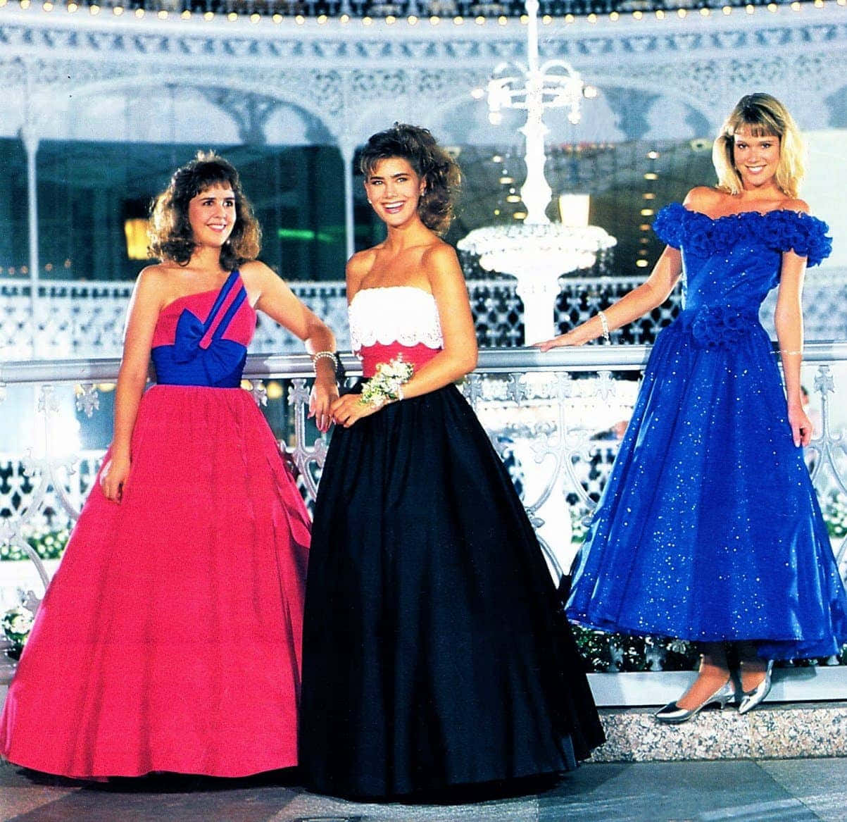 三位穿著多彩1980年代禮服的女性在夜晚的噴泉旁微笑。