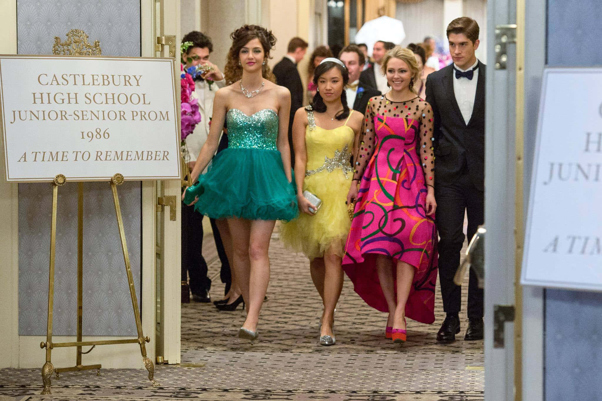 80年代舞會的璀璨之夜 壁紙 — 青少年穿著鮮豔的1986年舞會禮服和西裝，走過宏偉的走廊，經過Castlebury高中的初級高級舞會標誌。