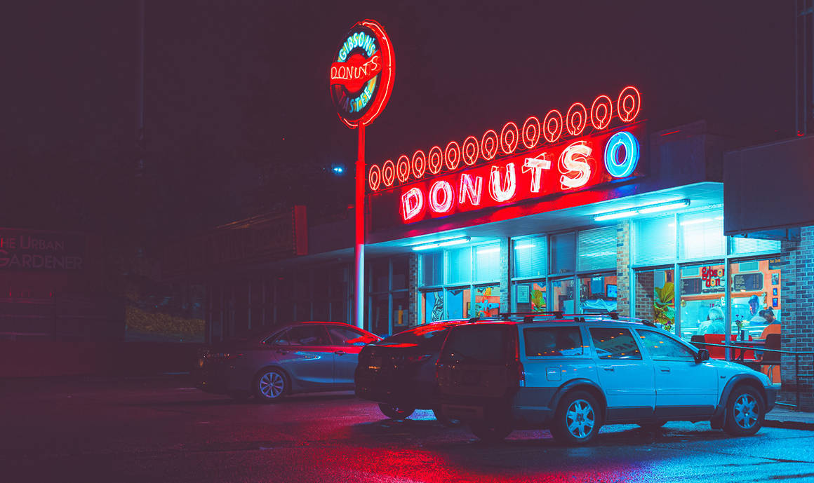 80年代風格霓虹甜甜圈店 iPhone 壁紙 — 夜晚一間霓虹燈光下的冬甩店，發光的紅色和藍色招牌倒映在濕漉漉的路面上，外面停著汽車。