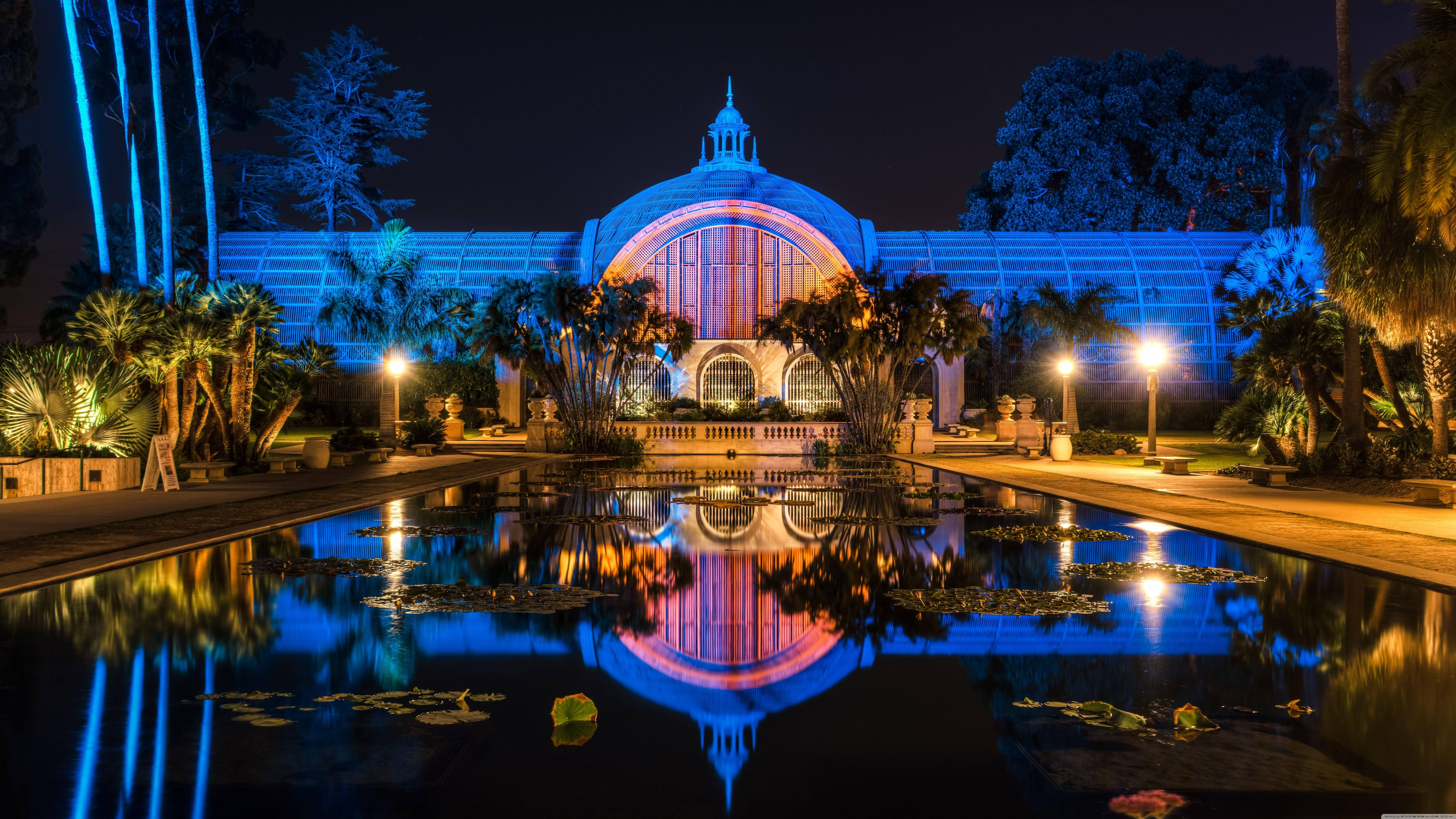 8K 7680x4320 超高清解析度桌面壁紙：加州聖地亞哥巴爾博亞公園夜景 壁紙 — 一個巨大的圓頂溫室在藍色和粉紅色燈光下照亮，完美地倒映在夜晚寧靜的池塘中，周圍環繞著棕櫚樹。