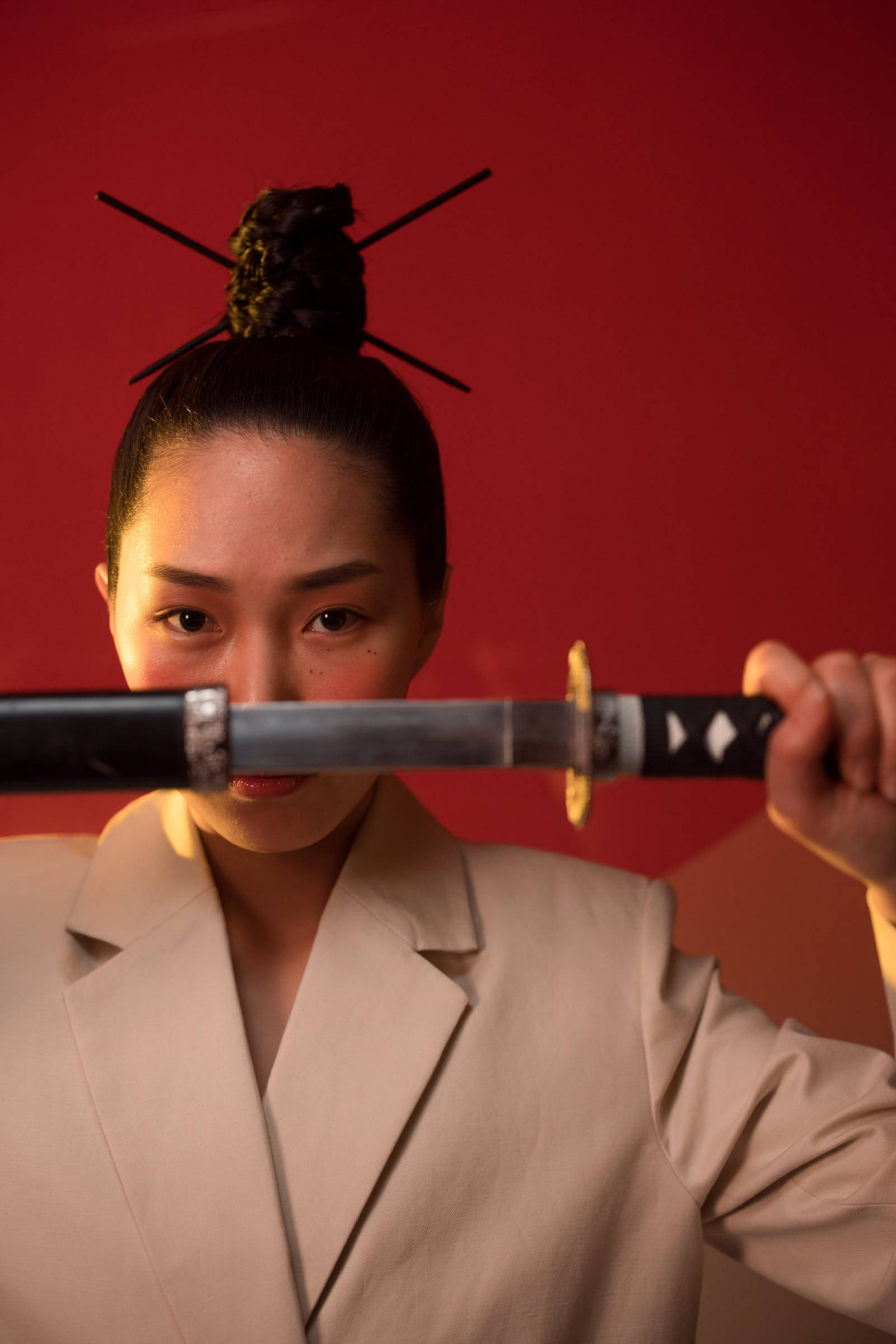 8K 武士女子持銀色薙刀 壁紙 — 一位身穿米色西裝的女性水平握著一把武士刀，她強烈的目光與觀眾對視，背景是鮮明的紅色。