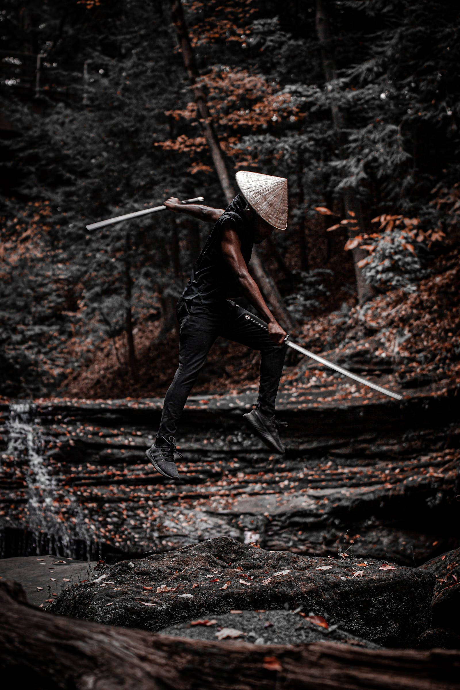 8K 武士揮劍於森林 壁紙 — 一位戴著圓錐帽的戰士在空中躍起，手持雙劍，下方是長滿苔蘚的森林溪流，散落著秋葉。