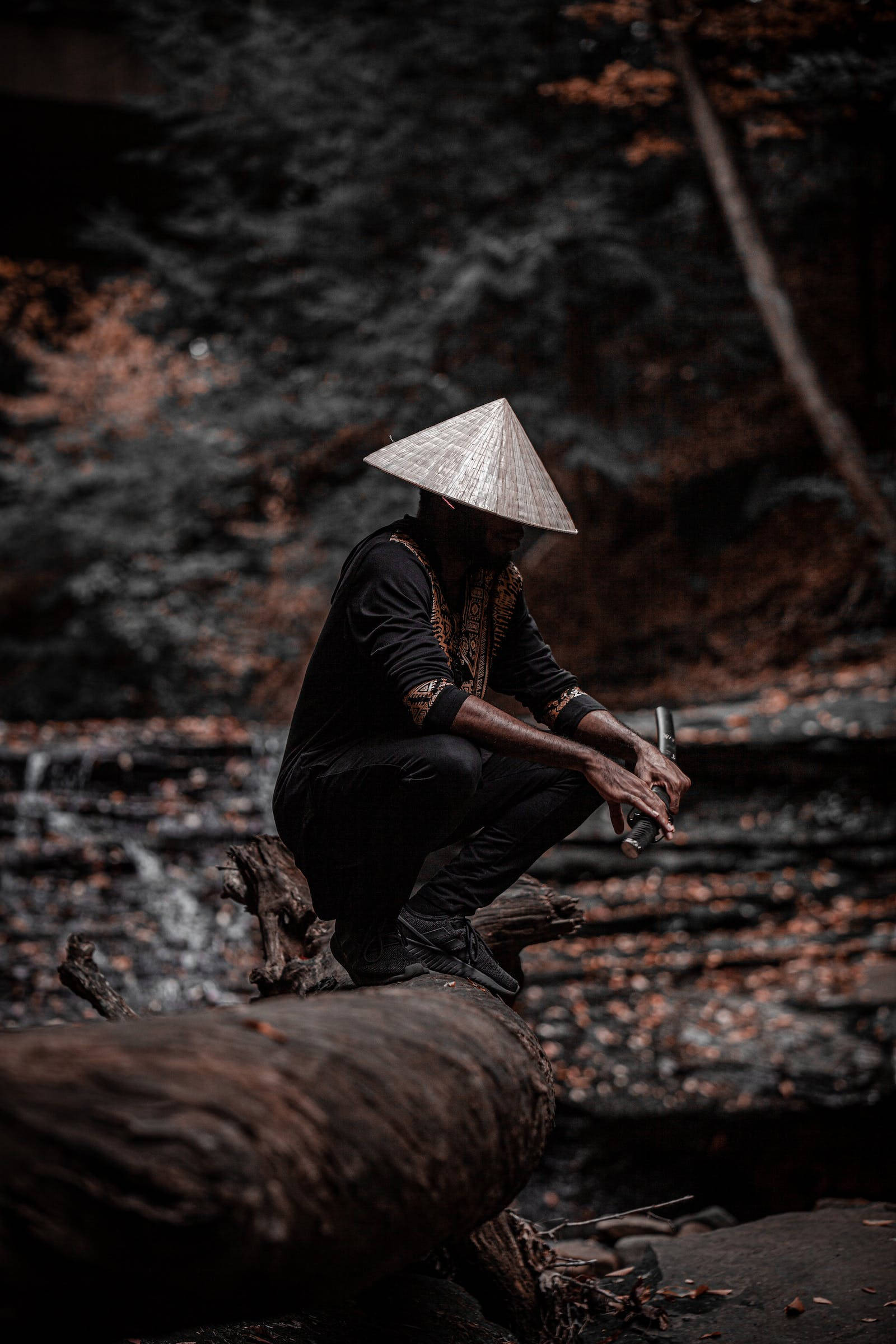 8K 武士暗黑風格 壁紙 — 一個戴著圓錐形帽子的人蹲在黑暗森林中一條長滿苔蘚的小溪旁邊的倒下的木頭上。
