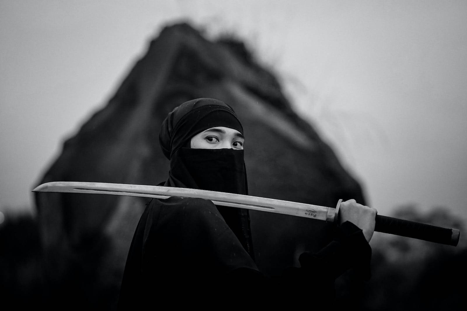 8K 武士肩扛薙刀 壁紙 — 一個戴面具的忍者手持武士刀，背景是模糊的山脈，呈現鮮明的黑白對比。
