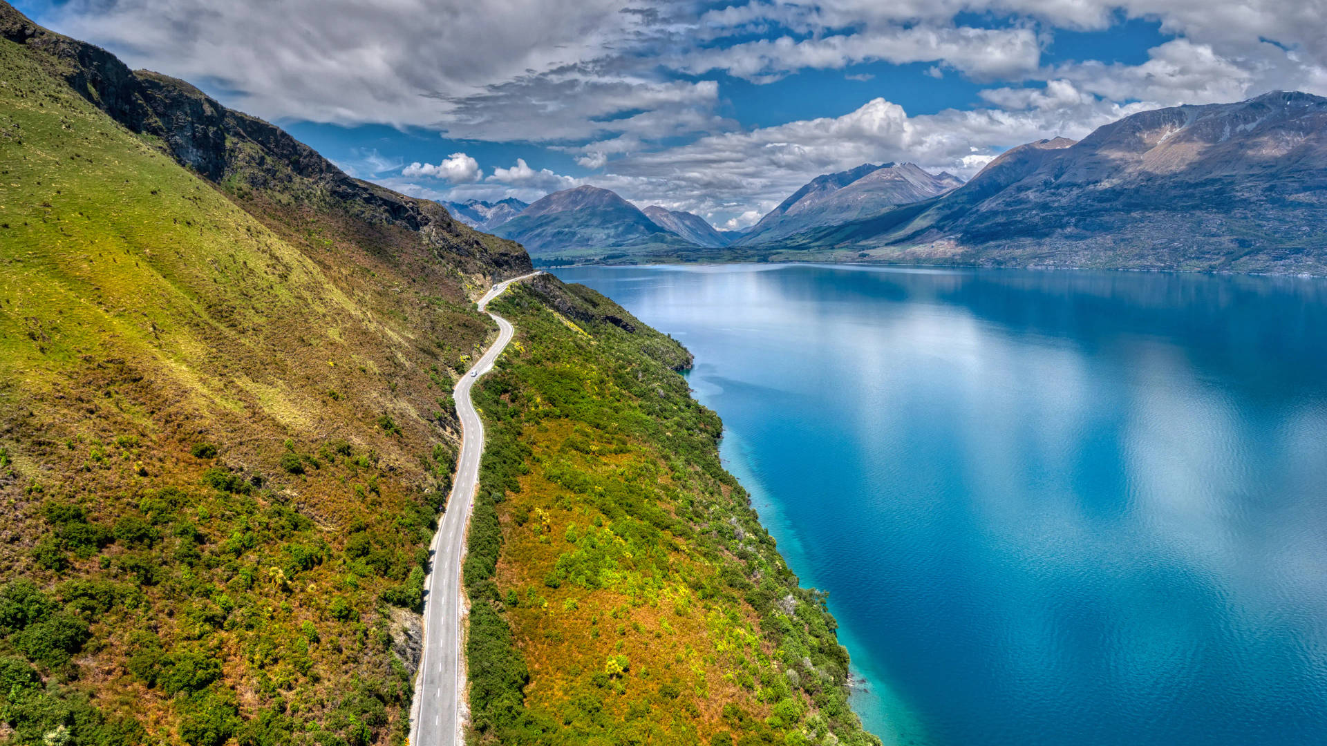 一條蜿蜒的道路沿著綠色山坡，旁邊是碧綠湖泊，背景有山脈。