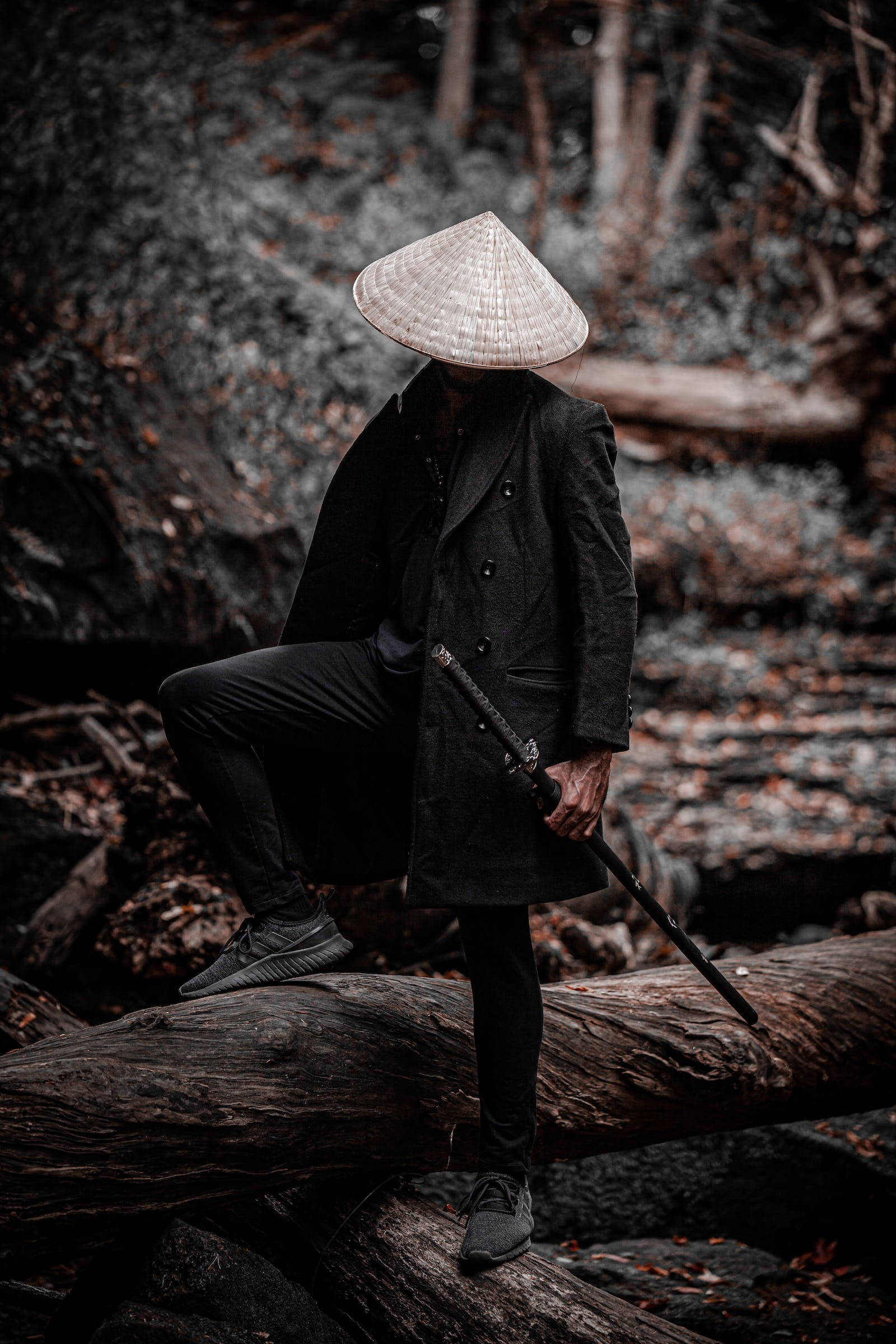 8K武士全黑裝束戴草帽 壁紙 — 一個身穿黑色外套和圓錐形帽子的人物坐在陰暗森林中的一根倒下的木頭上，手持一把帶有隱藏面孔的劍。