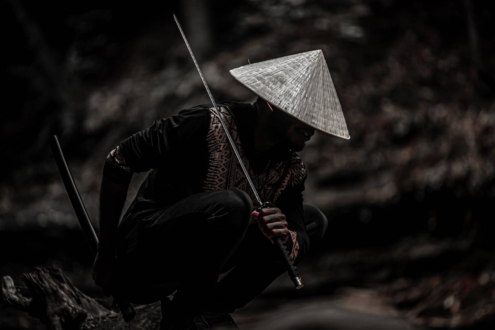 8K武士戴灰草帽持武士刀 壁紙 — 一位戴著圓錐帽的孤獨戰士蹲在陰暗的森林中，專注地握著劍。