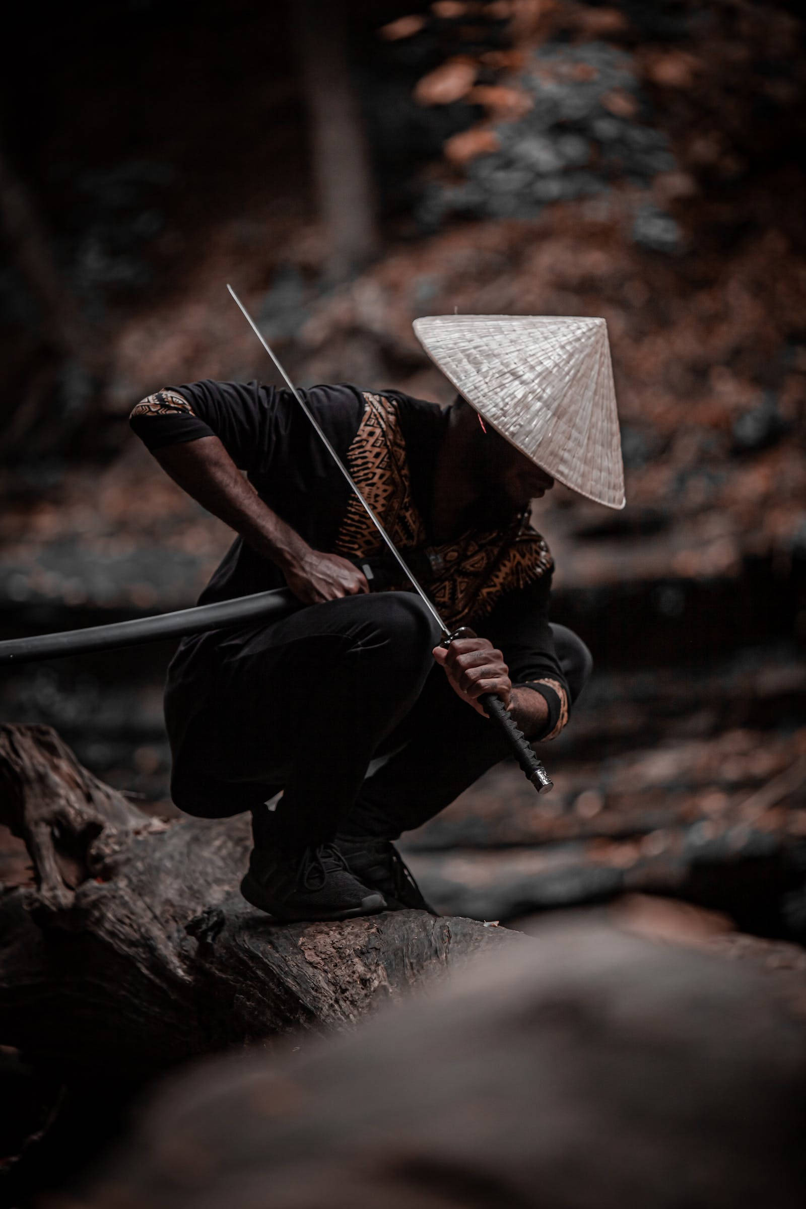 8K武士戴草帽 壁紙 — 一個戴著圓錐形帽子的人蹲在木頭上，在秋葉飄落的森林中手持長劍。