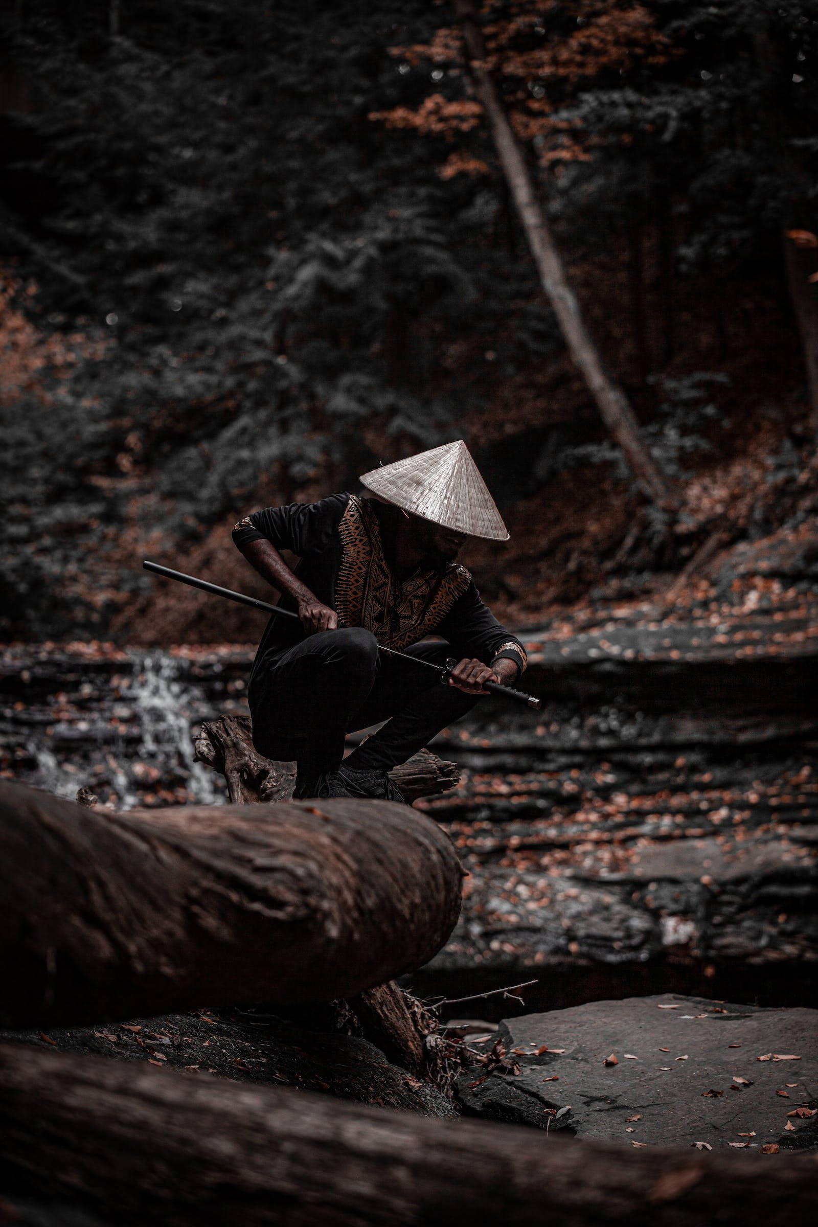 8K武士拔刀姿勢 壁紙 — 一個戴著圓錐形帽子的人蹲在森林溪流旁的圓木上，手持長杖。