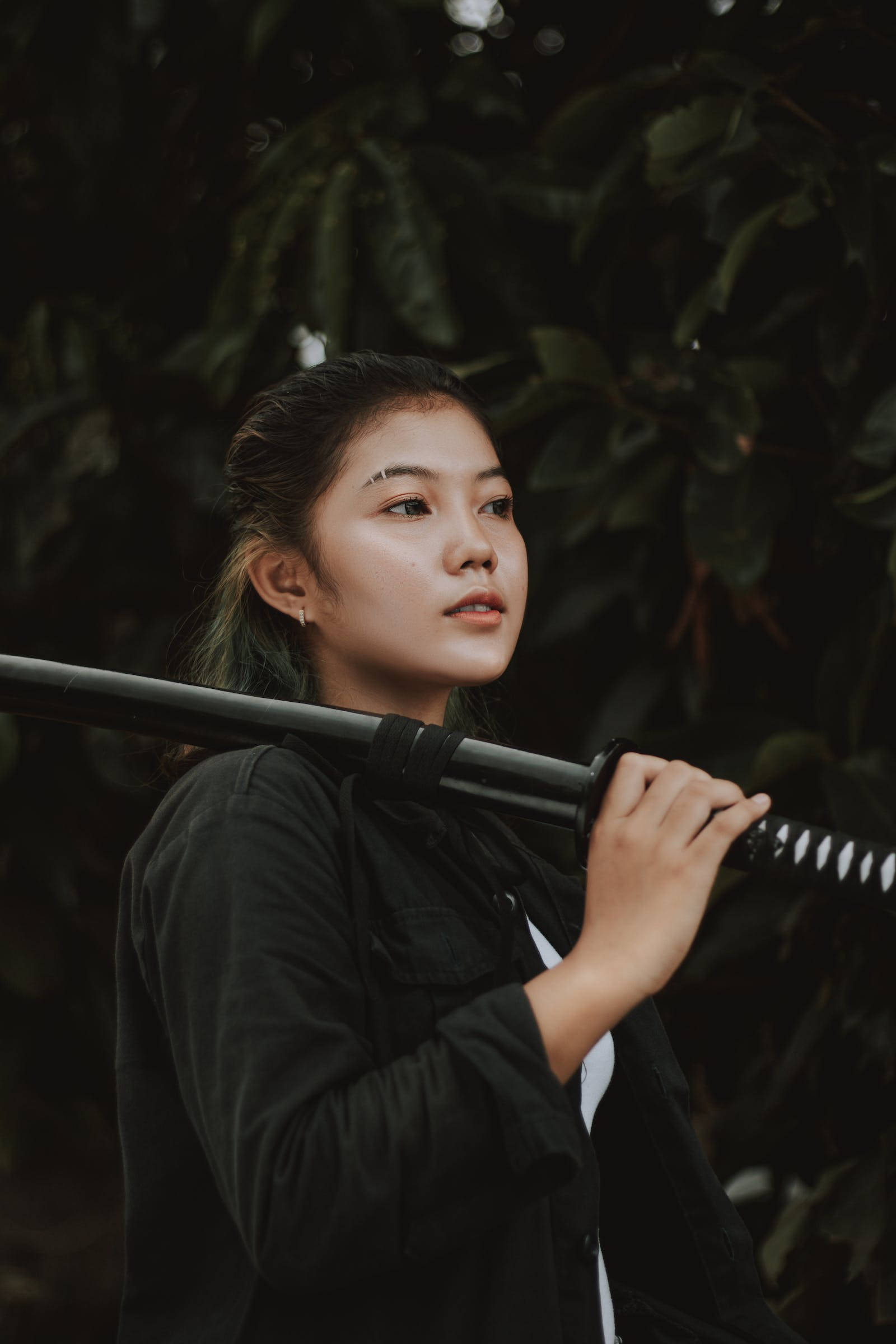 8K武士持黑色武士刀 壁紙 — 一位黑髮年輕女子在茂密綠葉背景下將武士刀扛在肩上。