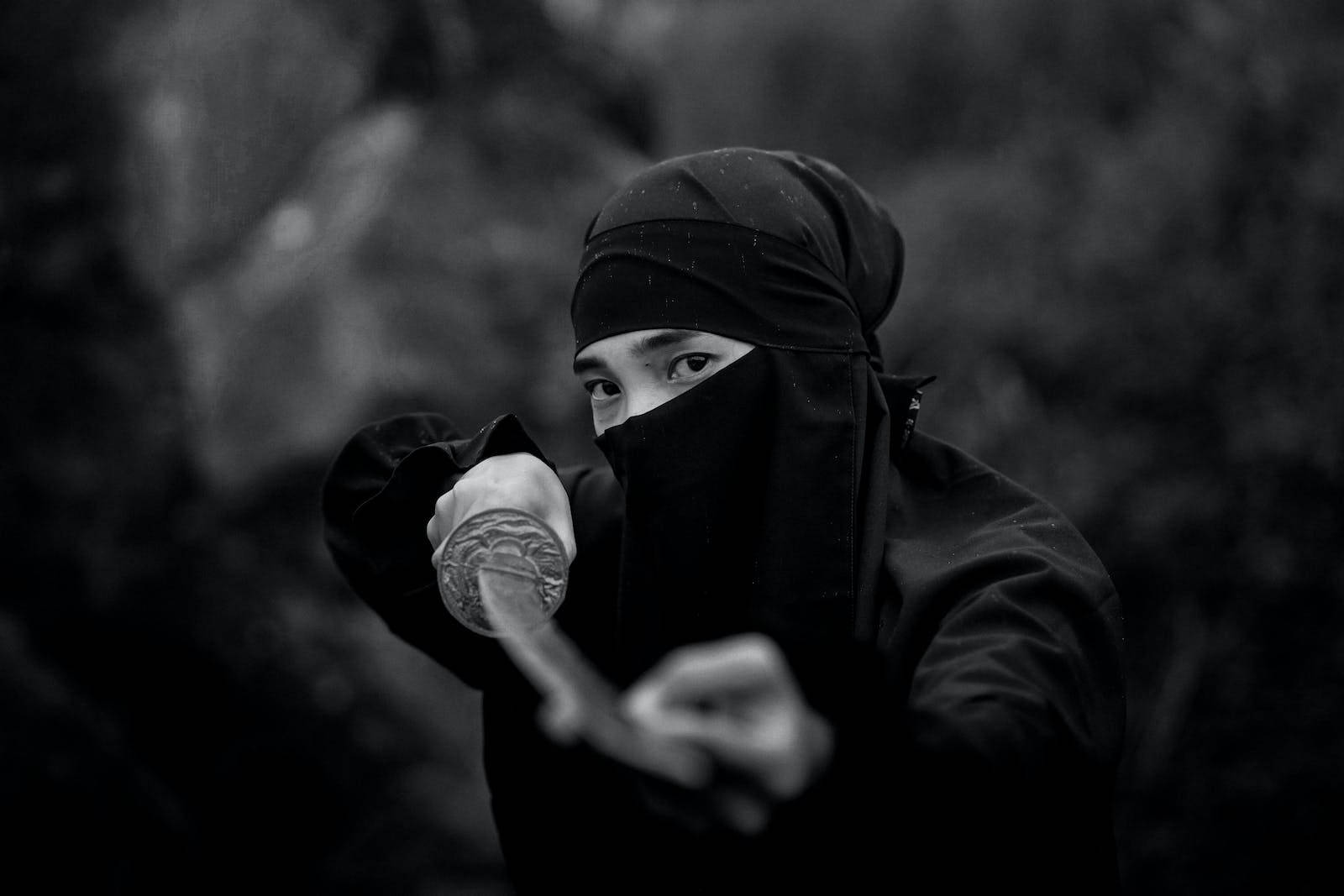 8K武士穿黑色忍者服 壁紙 — 一個戴黑色面具的忍者，在森林中手持武士刀，眼神專注而強烈，以單色呈現。