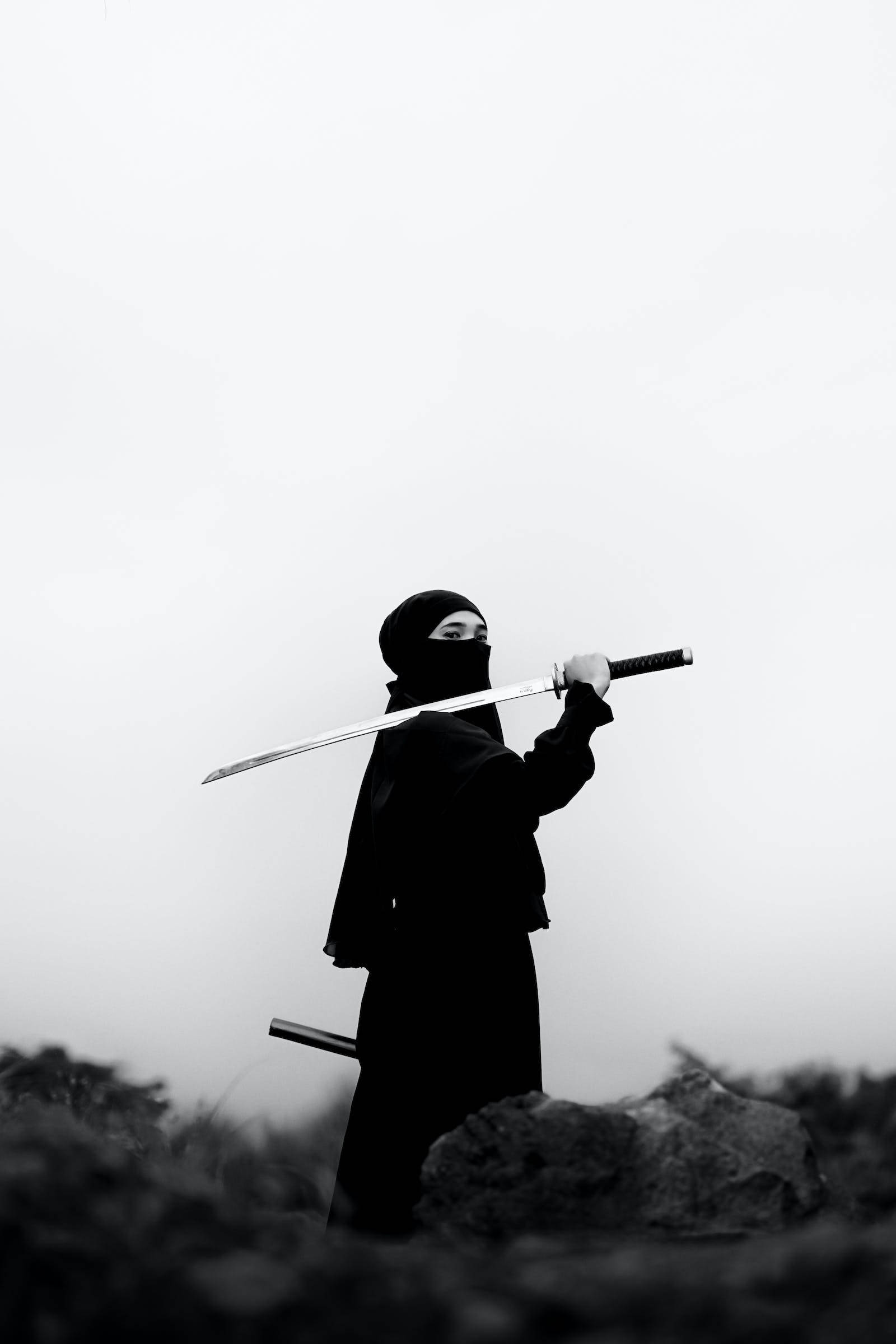 8K武士穿黑色服裝 壁紙 — 一名身穿黑色裝備的忍者，在霧氣瀰漫的灰色天空下手持武士刀，下方有岩石。