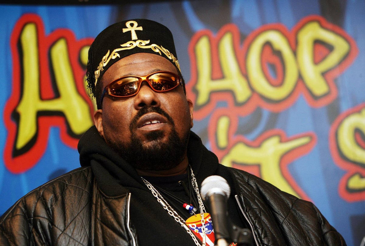 90年代饒舌歌手Afrika Bambaataa 壁紙 — 一名戴著帶有金色安卡和太陽眼鏡的黑色帽子的男子，在充滿活力的藍色背景前，對著麥克風說話，背景上有黃色塗鴉風格的「HIP HOP」字樣。