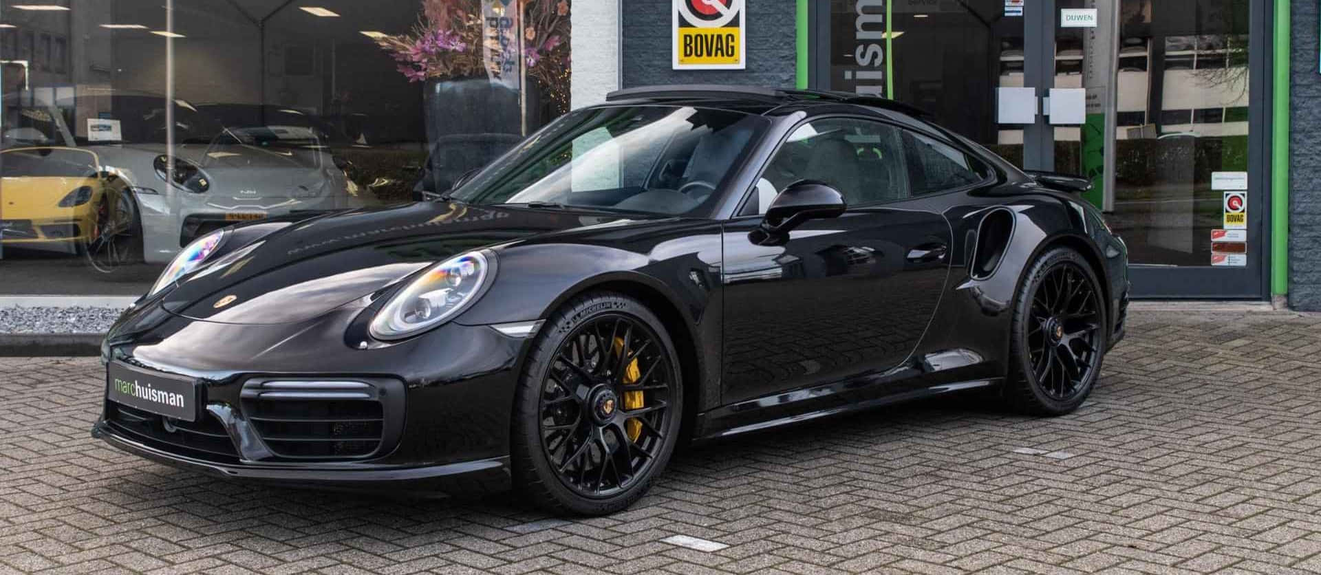 黑色保時捷911 – 4K汽車壁紙 壁紙 — 一輛型格黑色保時捷911 Turbo S，配備黃色制動卡鉗，停在汽車經銷商前的磚砌車道上。