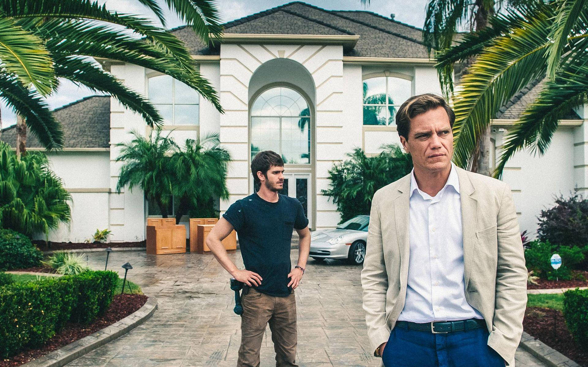 《99 Homes》電影海報 壁紙 — 兩個男人站在石徑上，面前是一座宏偉的白色豪宅，周圍環繞著棕櫚樹和茂密的綠植，天空多雲。