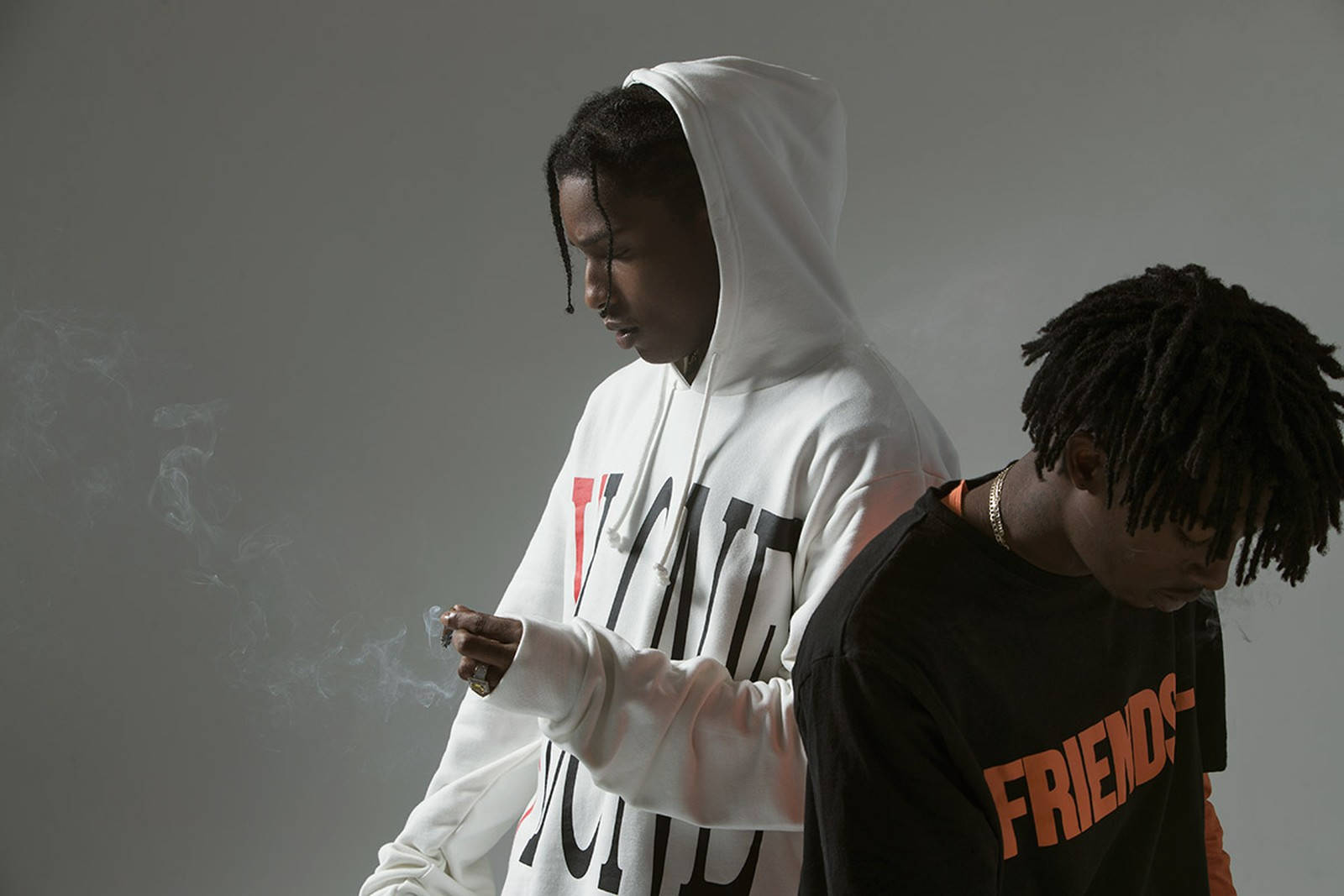 A$AP Rocky 與 A$AP Bari VLONE 壁紙 — 兩個穿連帽衫的年輕男子靠著灰色牆壁站立，一人抽煙，另一人低頭，兩人均表情嚴肅。