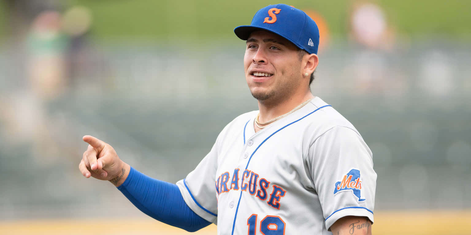  壁紙 — 一名身穿 Syracuse Mets 球衣的棒球運動員在陽光明媚的場地上進行日間比賽時微笑並指向某處。