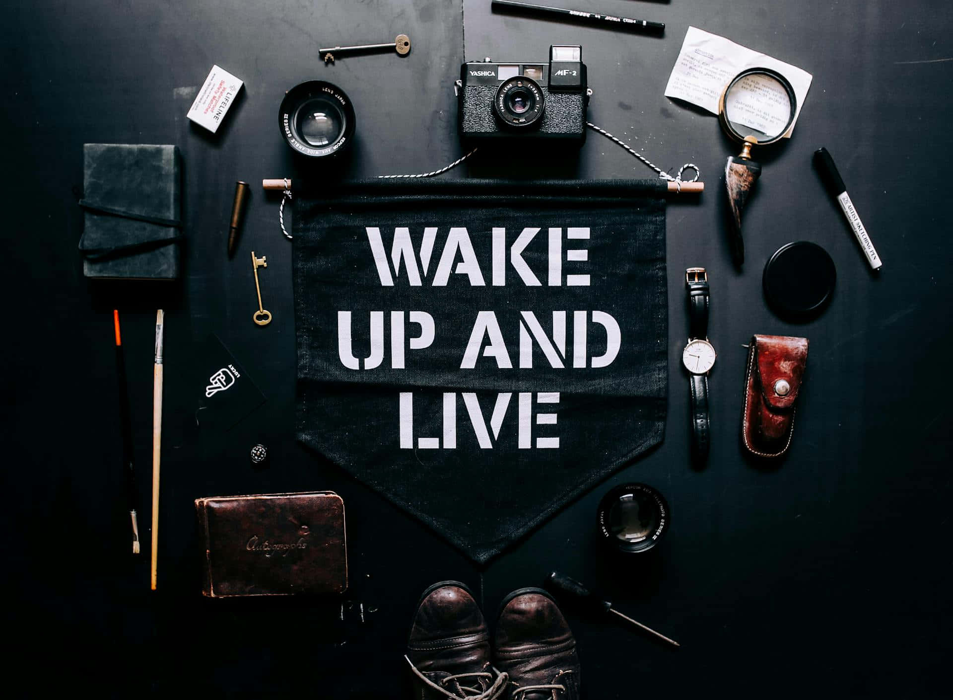  壁紙 — 一個黑色橫幅，上面寫著「WAKE UP AND LIVE」，周圍環繞著復古相機鏡頭、筆記本和皮革製品，放在深色表面上。