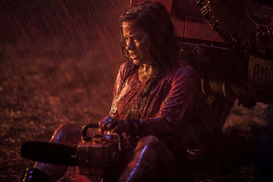  壁紙 — 一個血淋淋的女人在雨浸的田野中，在昏暗的紅光下緊握著電鋸。