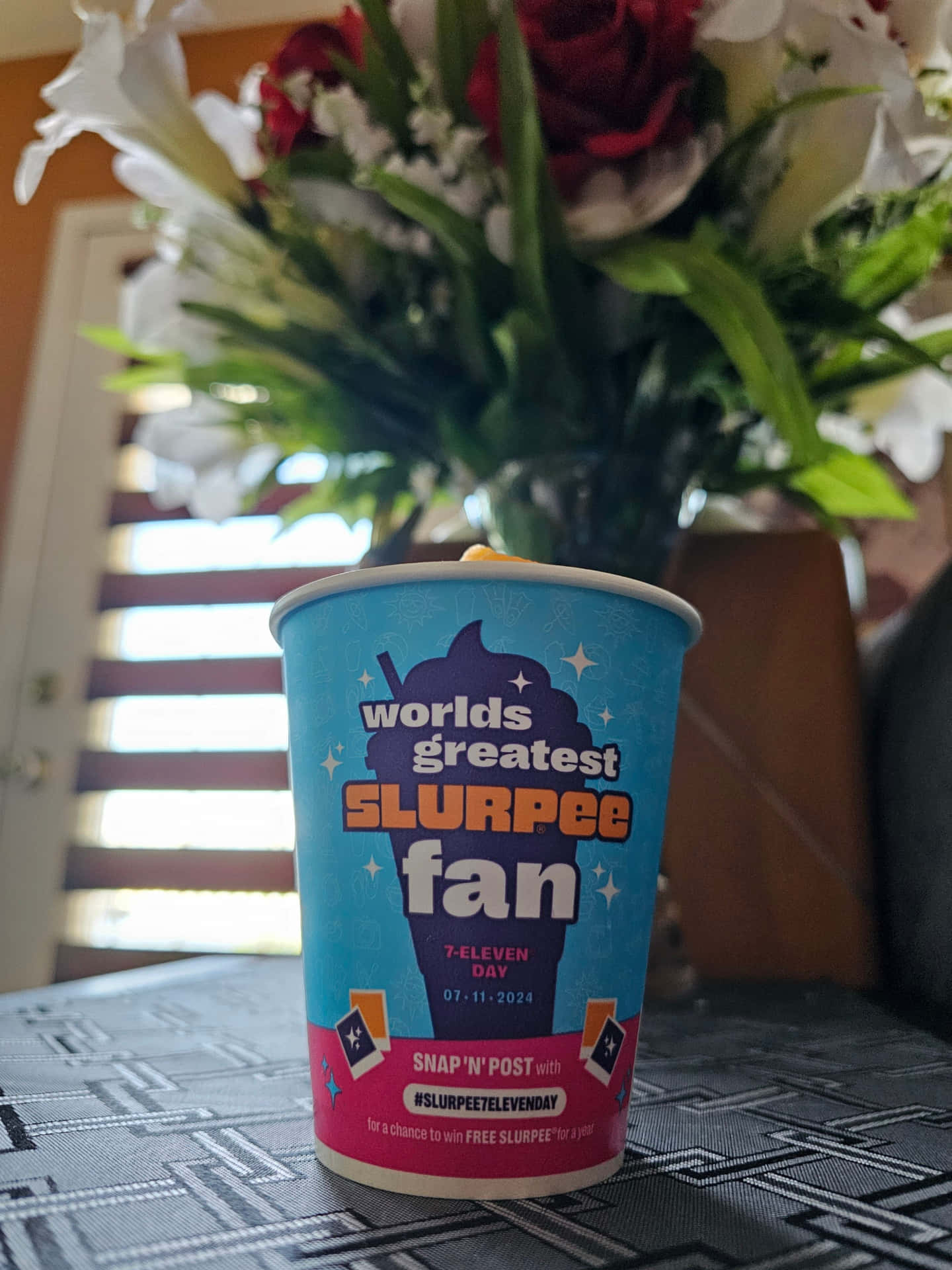  壁紙 — 一個藍色和粉紅色的7-Eleven Slurpee杯，標有「worlds greatest SLURPEE fan」，放在一張有圖案的桌子上，背景有一束紅色和白色的花。