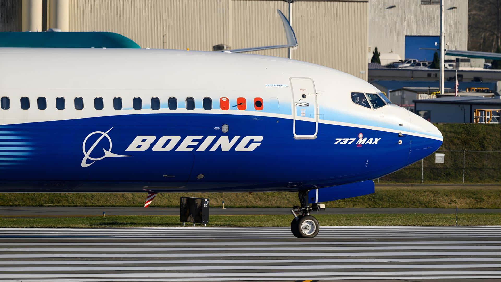  壁紙 — 一架波音737 MAX飛機停在有條紋的停機坪上，背景是工業機庫，在明亮的日光下。