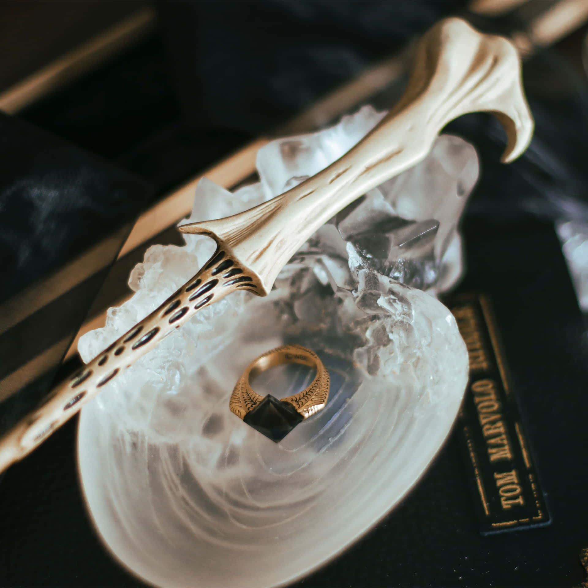  壁紙 — 一根類似骨頭的魔杖和一個金戒指放在一個水晶碗裡，霧氣從深色布料上升起，附近有一個名牌。