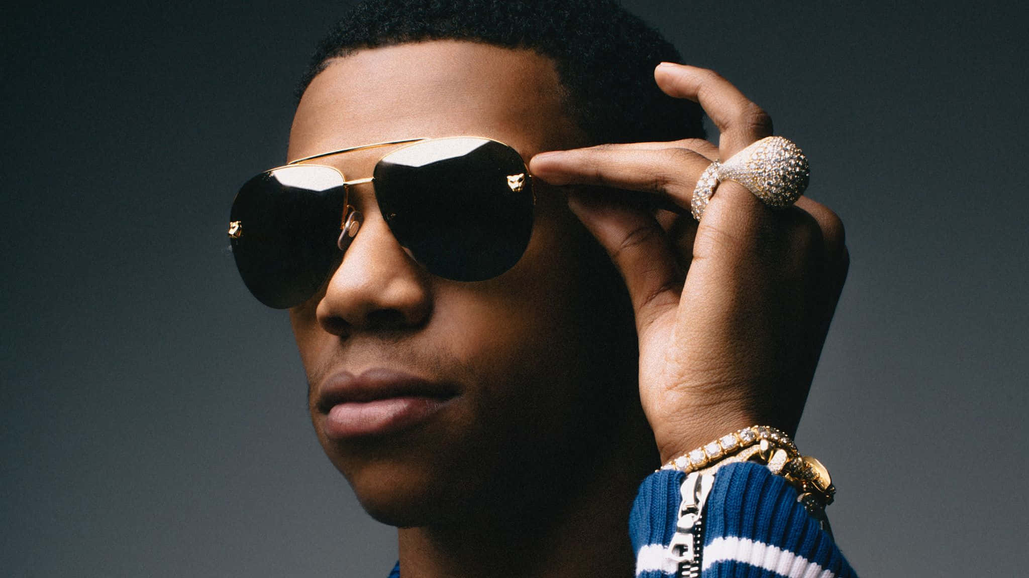 A Boogie Wit Da Hoodie 高光墨鏡 壁紙 — 一位時尚男士戴著飛行員太陽眼鏡，用戴著珠寶戒指的手調整眼鏡，背景為深灰色。