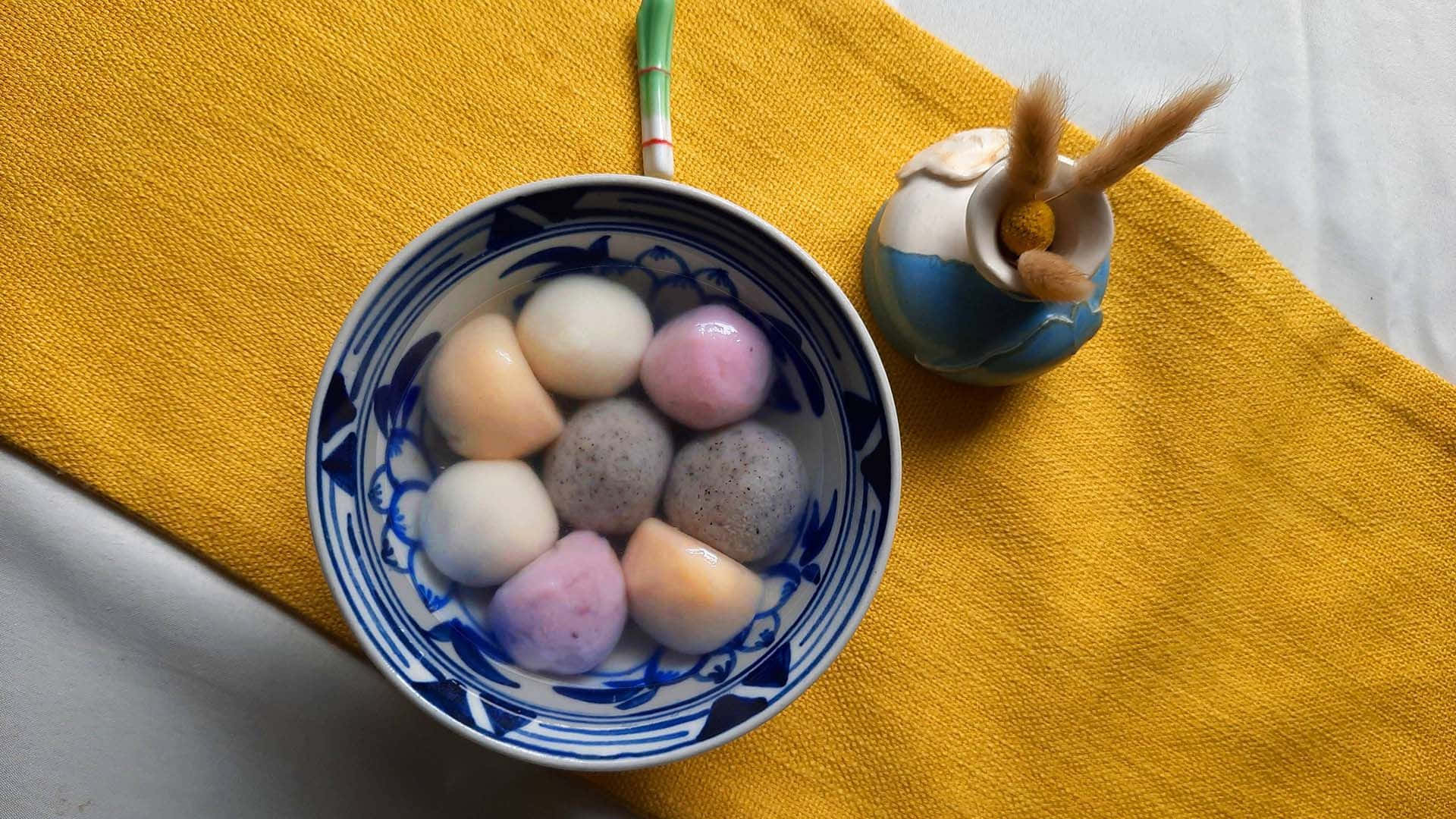  壁紙 — 一碗色彩繽紛的麻糬糰子放在藍白圖案陶瓷碗中，置於鮮黃色布上，旁邊有一個小裝飾罐。