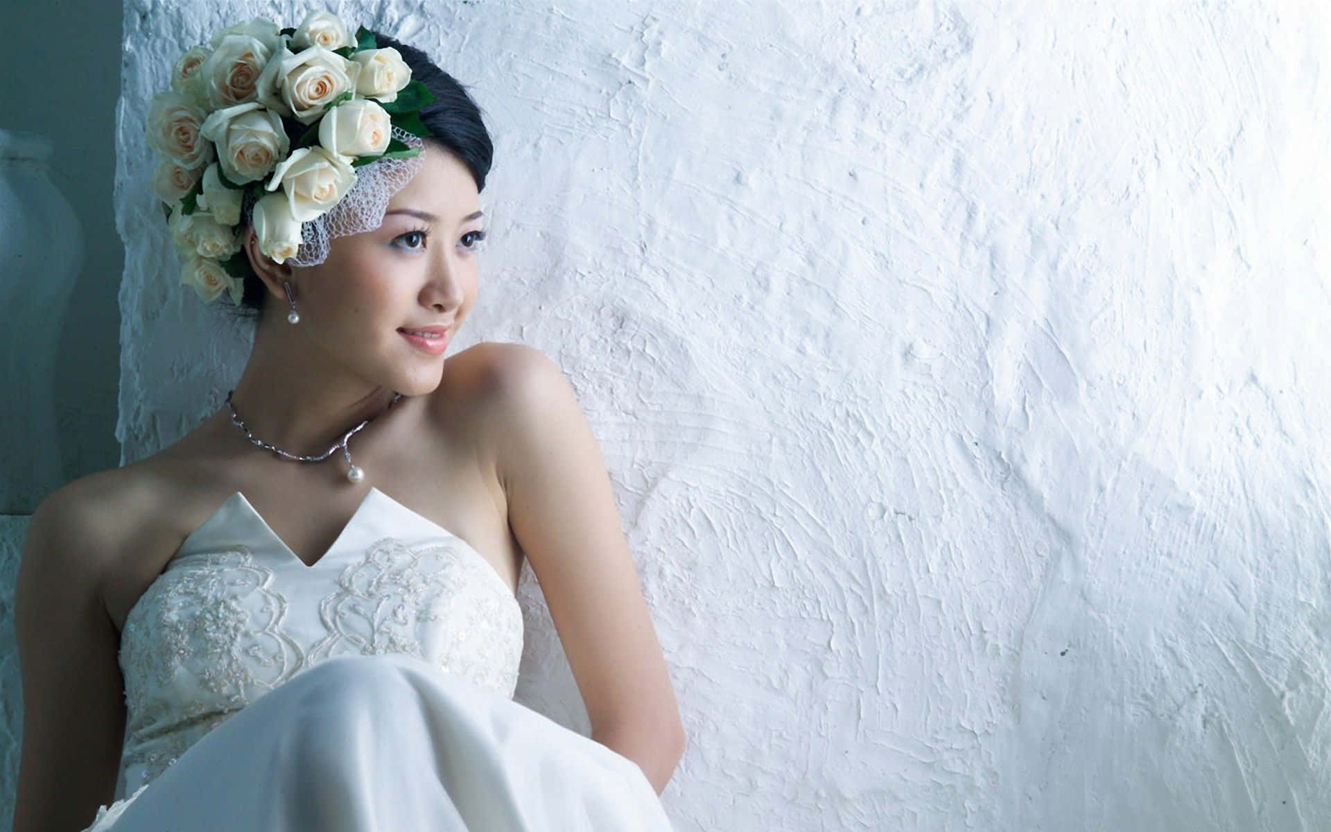 一位身穿白色蕾絲婚紗、頭戴玫瑰頭飾的新娘，對著白色牆壁微笑。