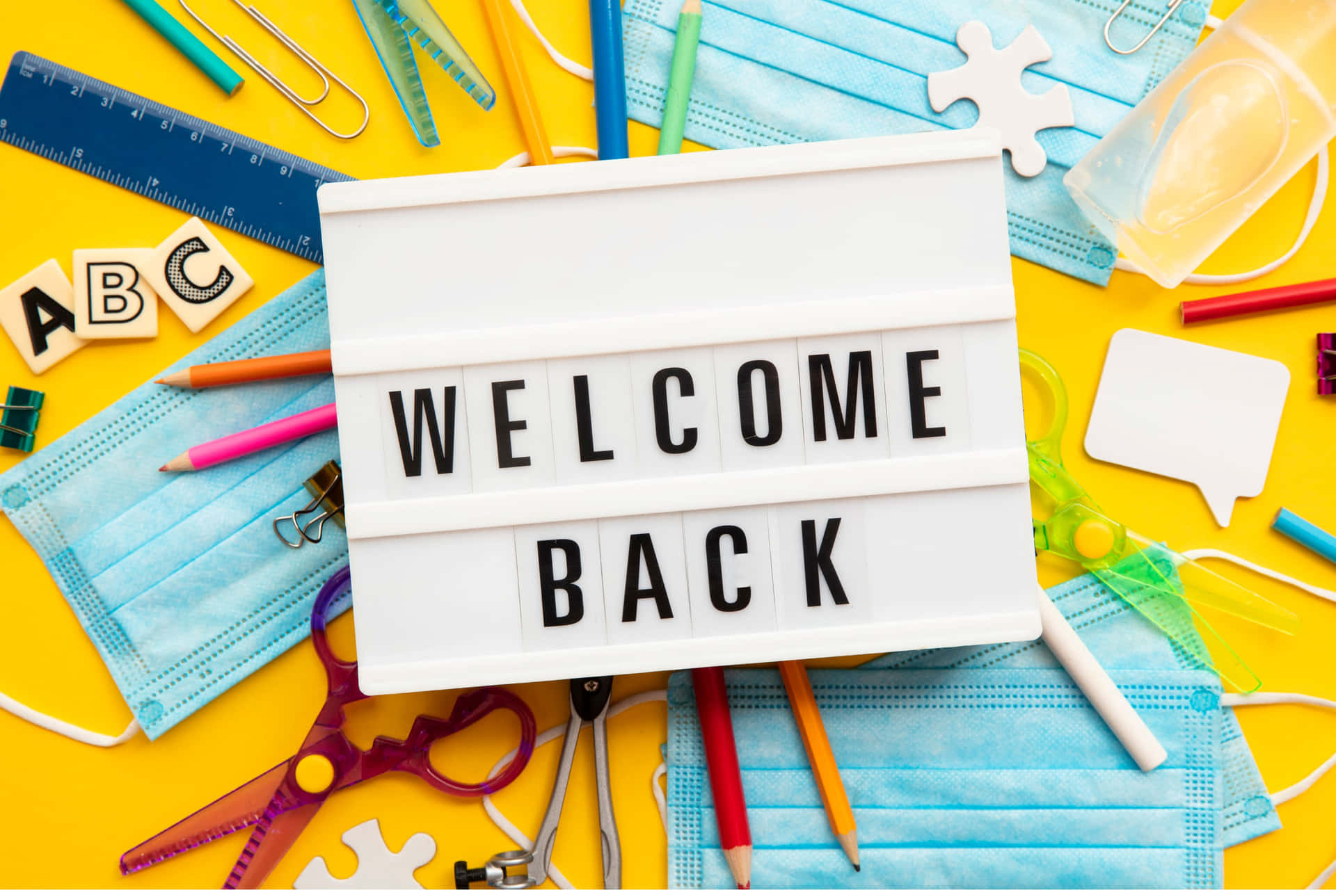  壁紙 — 一個明亮的黃色表面上展示了一個「WELCOME BACK」燈箱，周圍環繞著學校用品和藍色外科口罩。