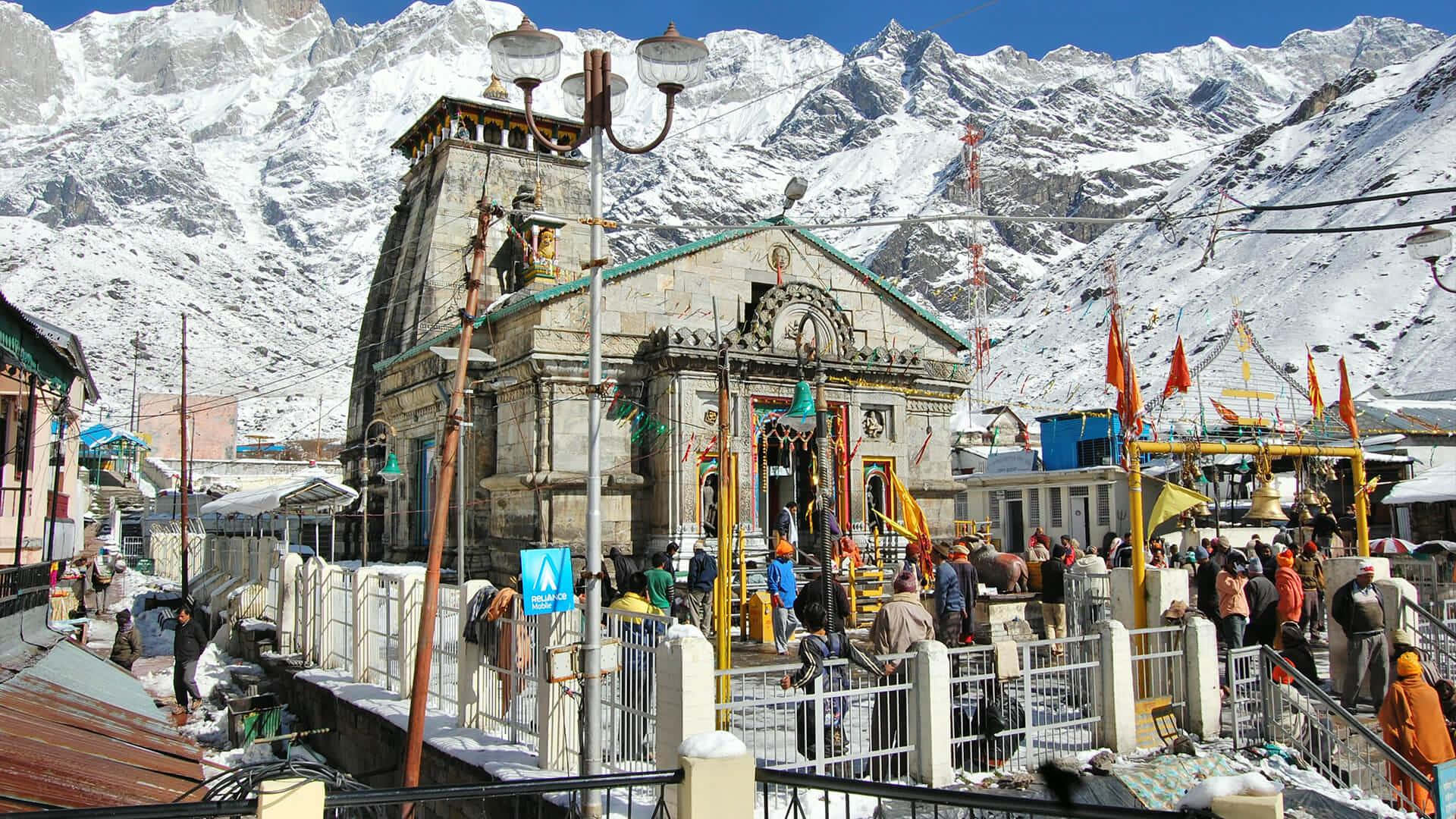 Kedarnath 桌布 壁紙 — 一座熱鬧的雪山寺廟，朝聖者聚集在其華麗的石結構周圍，背景是高聳的雪峰。