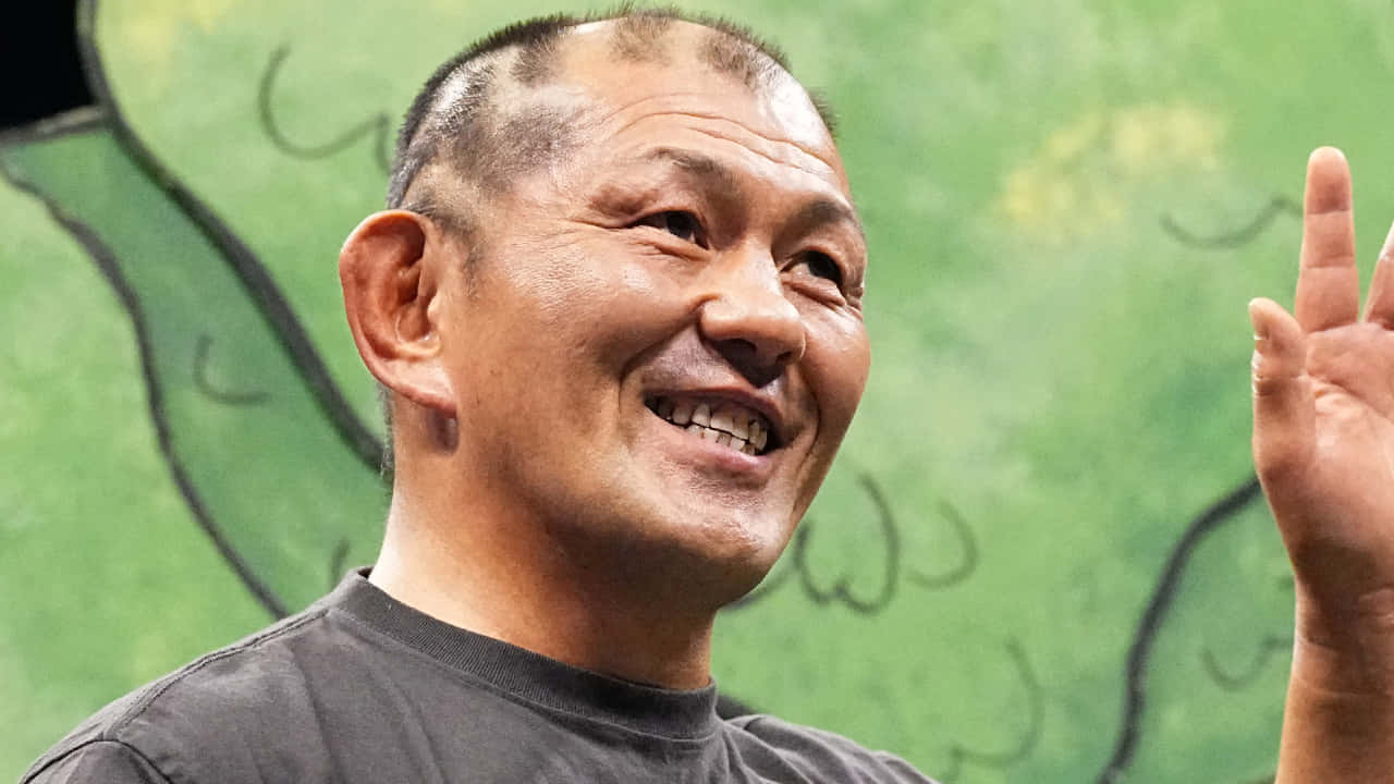  壁紙 — 一個光頭開朗的男子在綠色塗漆背景前微笑揮手。