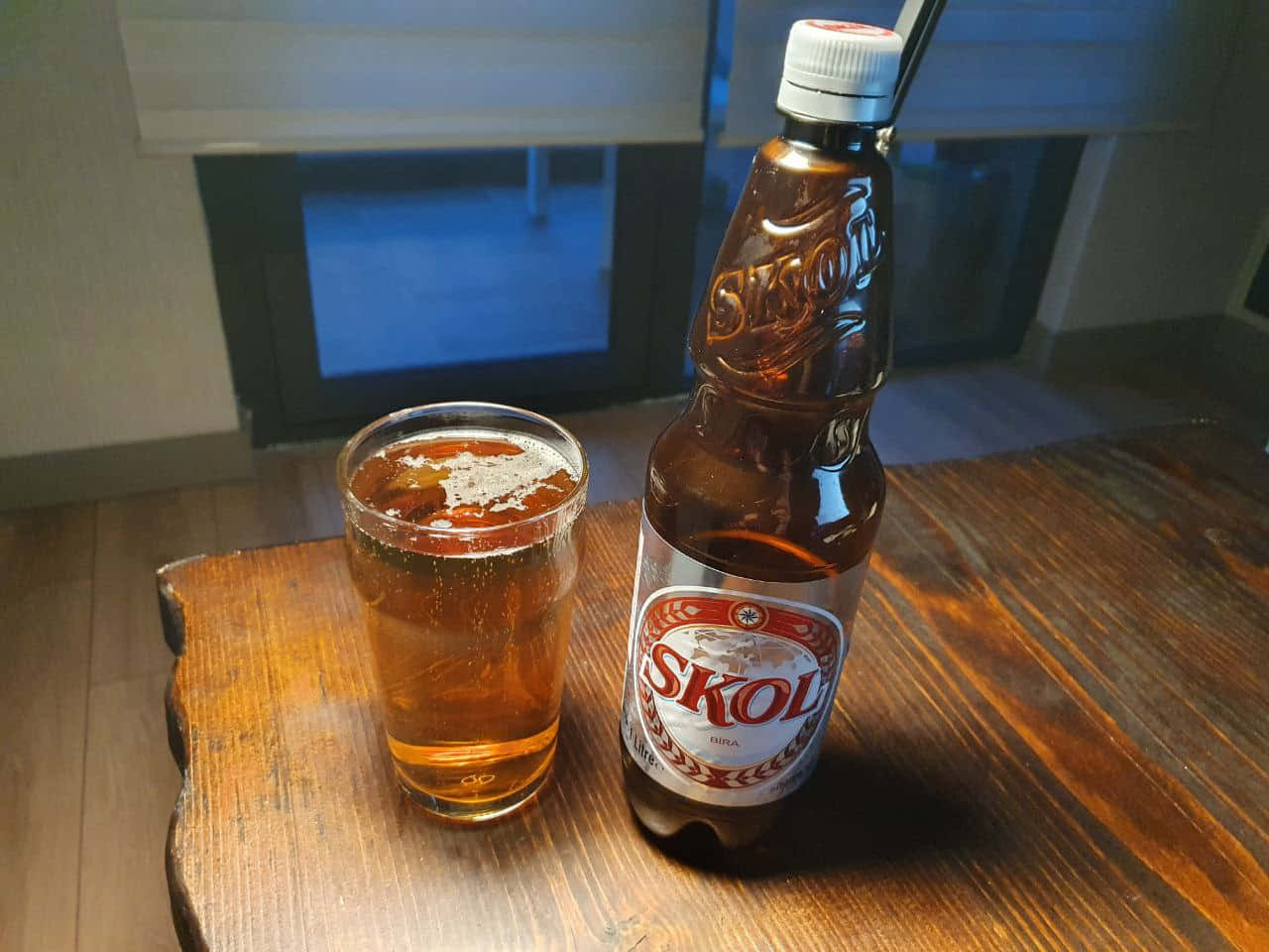  壁紙 — 一瓶冰凍的Skol啤酒和酒杯放在靠近藍色玻璃窗的質樸木桌上。