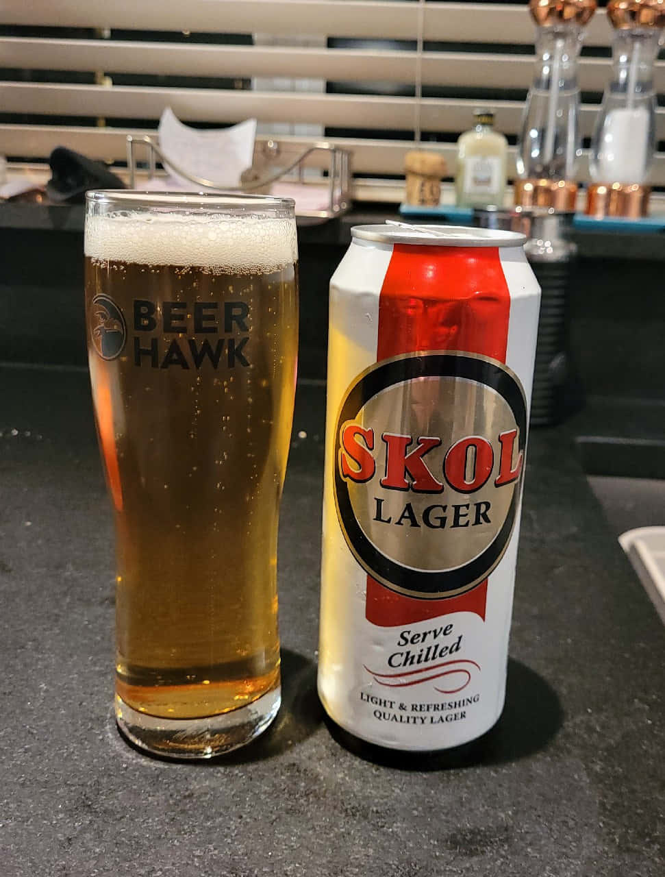  壁紙 — 一罐冰凍的Skol Lager啤酒放在室內燈光下的深色枱面上，旁邊有一個泡沫啤酒杯。