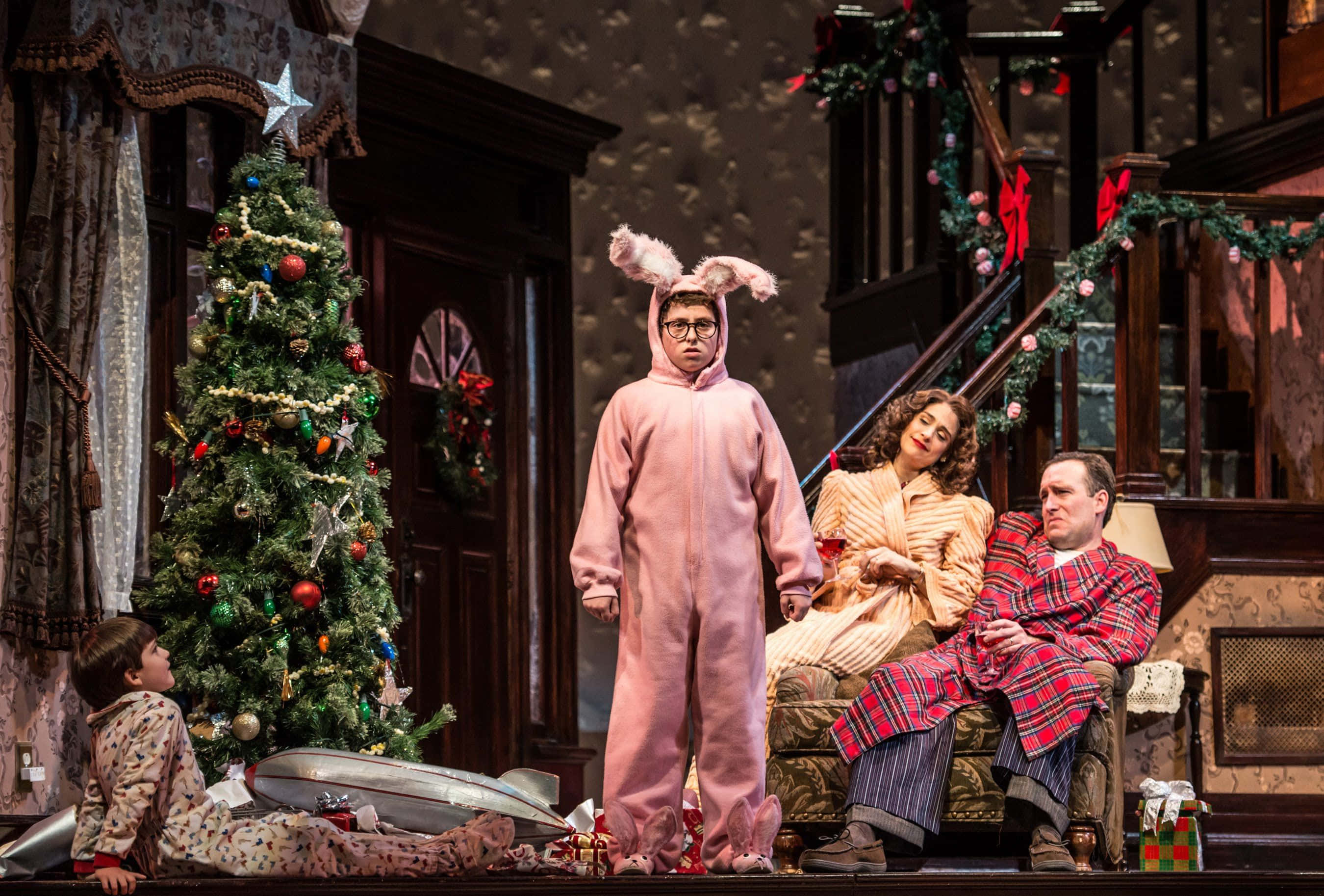 《A Christmas Story》音樂劇家庭場景 壁紙 — 一個穿著睡衣和節日服裝的家庭在溫暖照明的復古客廳中聚集在裝飾好的聖誕樹周圍。