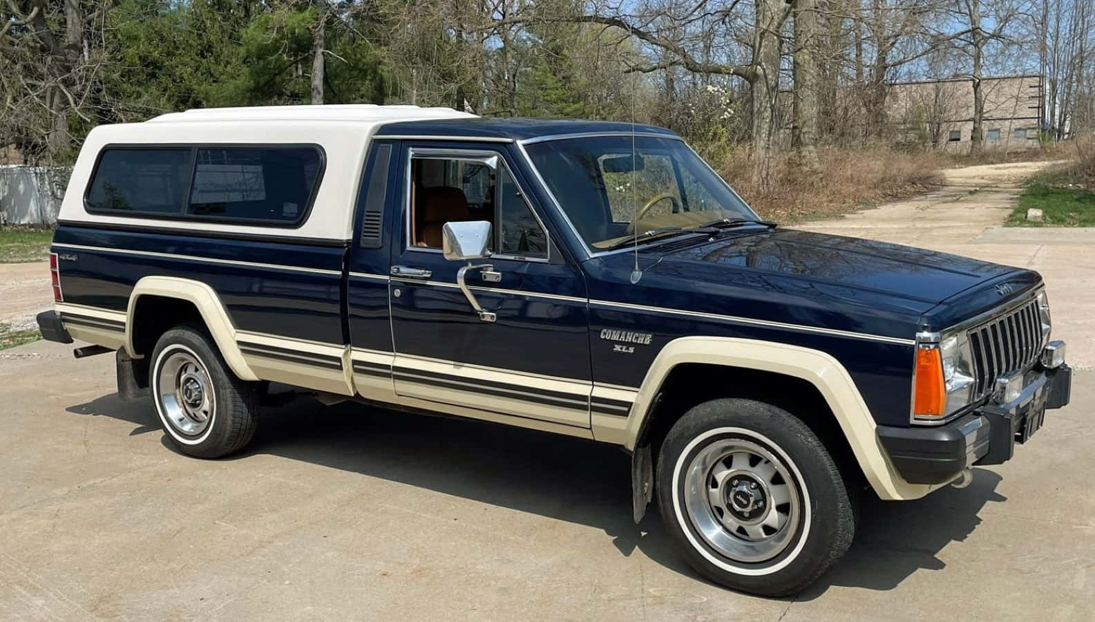 Jeep Comanche 桌布 壁紙 — 一輛經典藍白色Jeep Comanche皮卡車停在混凝土車道上，背景有樹木和建築物。